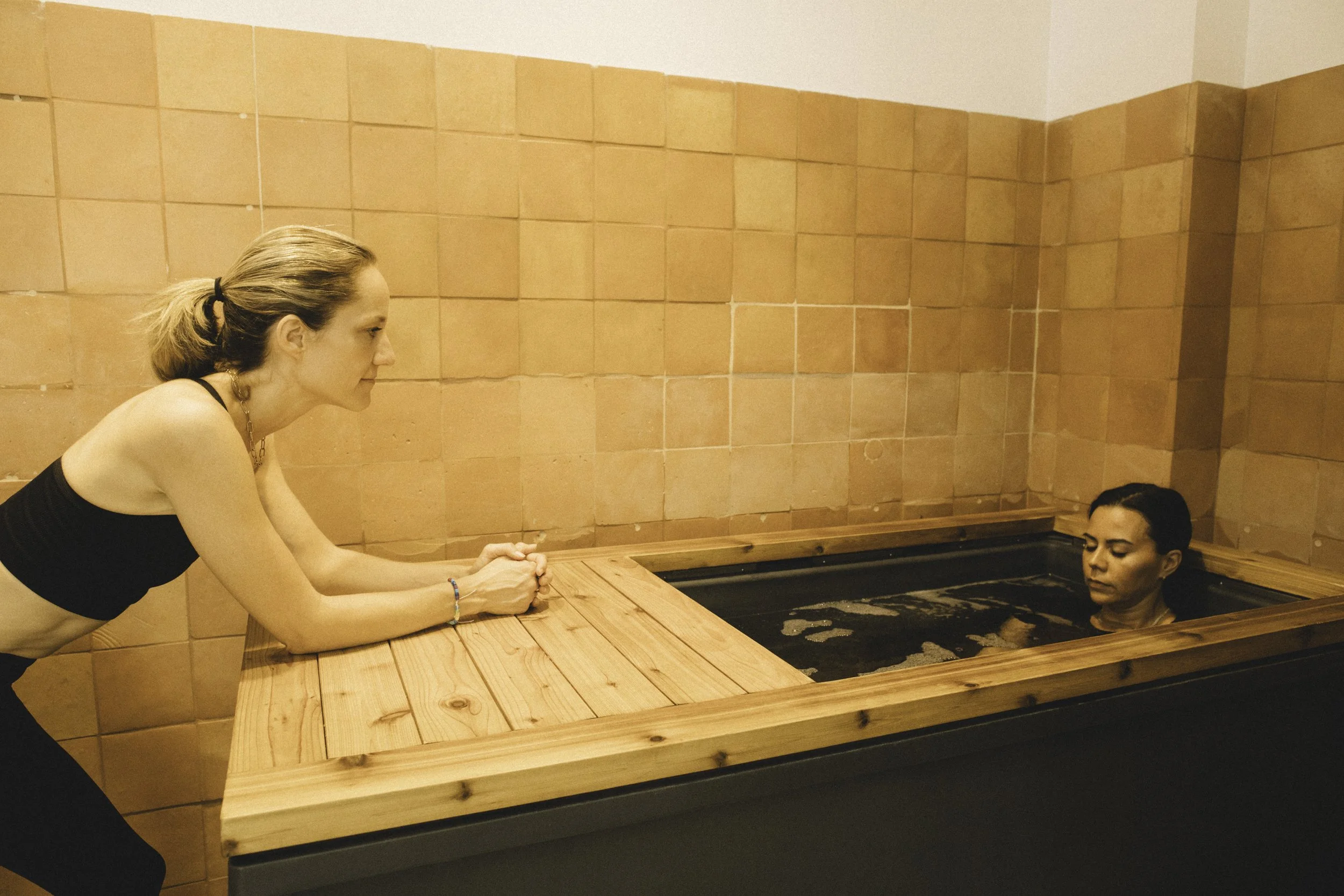 Two women in a spa, one sitting on the edge of a hot tub, the other submerged in the hot tub with only her head visible.