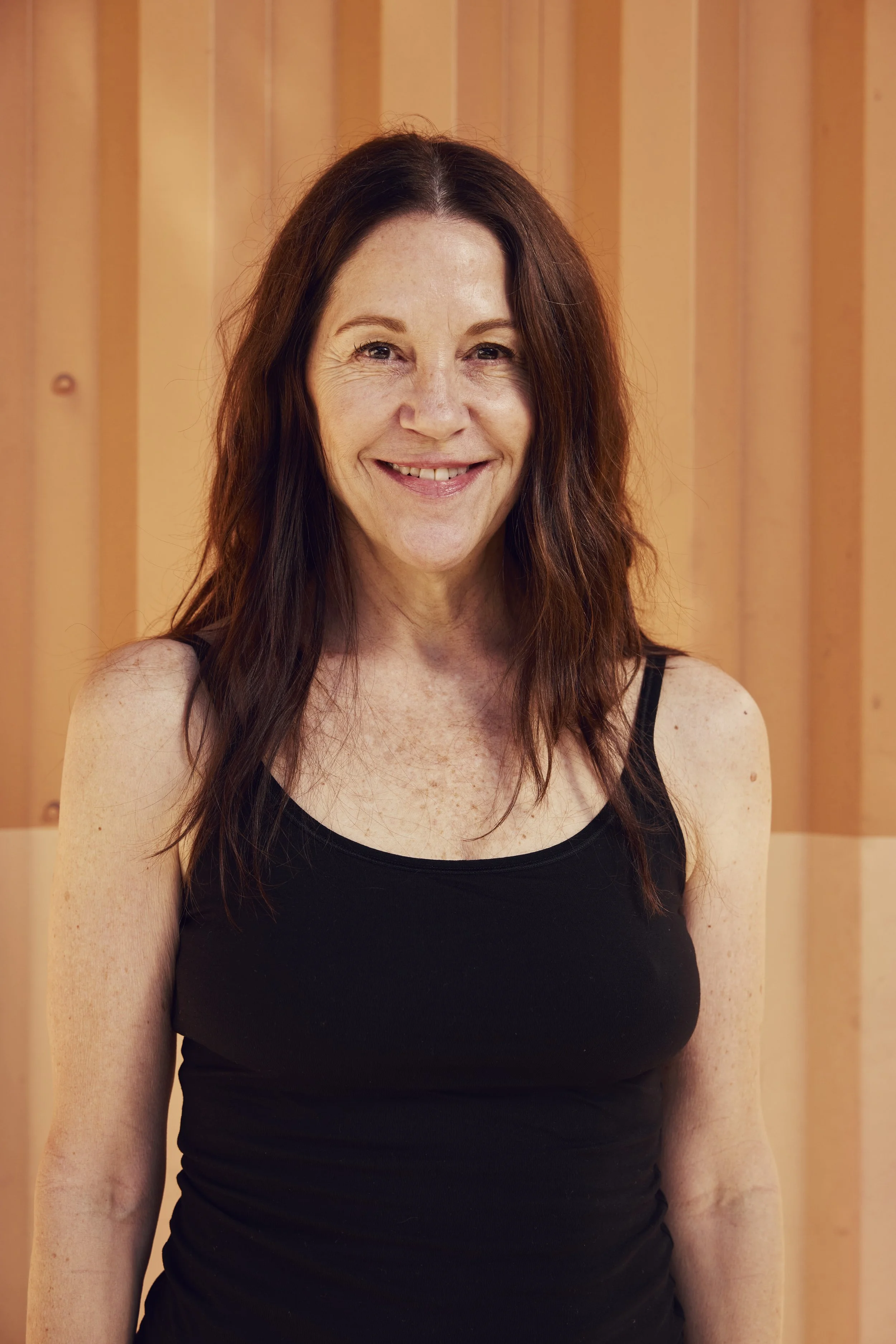 A woman with brown hair smiling and resting her arms on a table in an indoor setting with a blue poster or sign in the background and a green plant to the right.