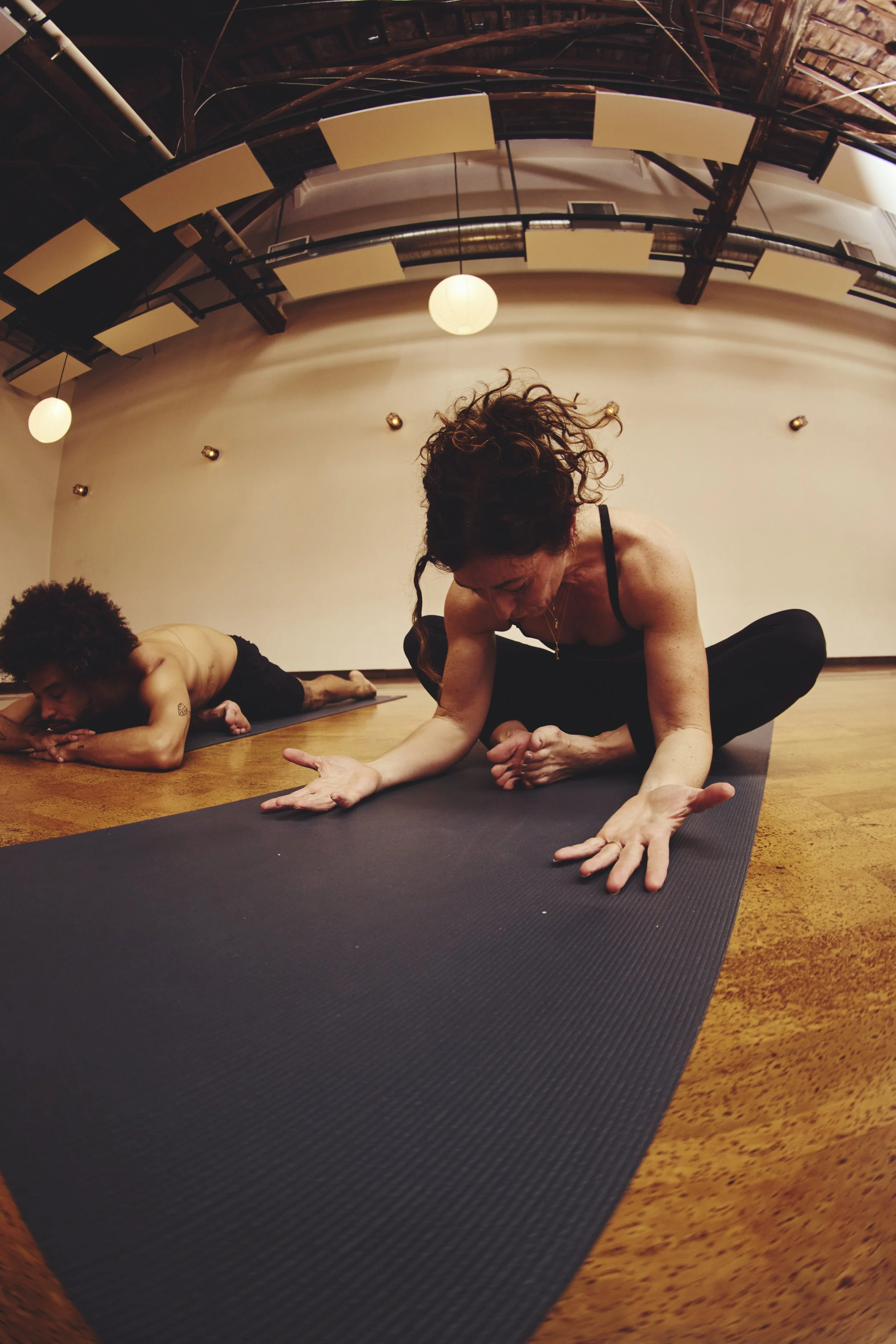 Two women practicing yoga on black mats in a spacious studio with warm lighting and wooden floors.