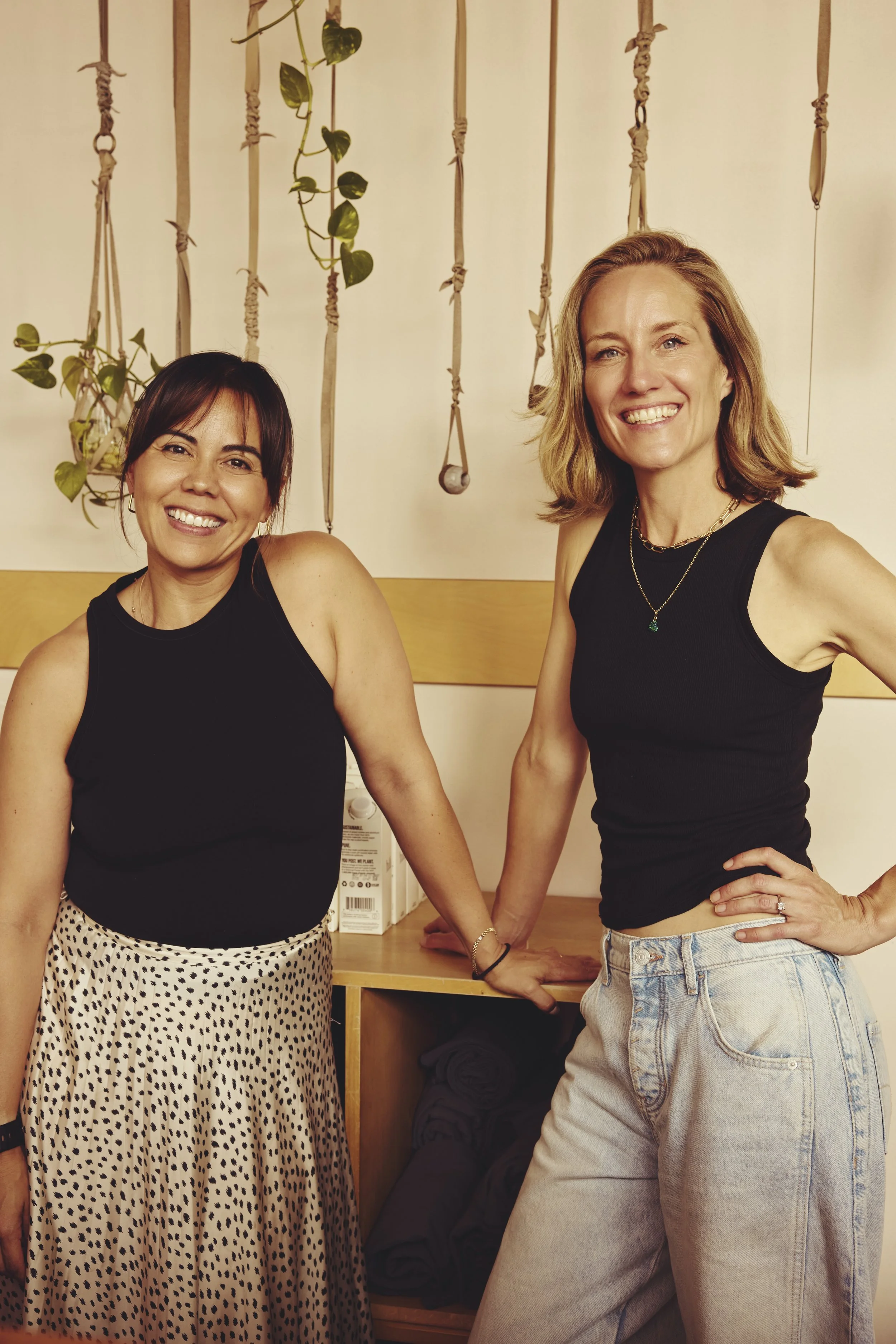 Two women smiling and posing for a photo in a room decorated with hanging plant and knotted ropes, one wearing a black sleeveless top and patterned skirt, the other in a black top and light-colored jeans.