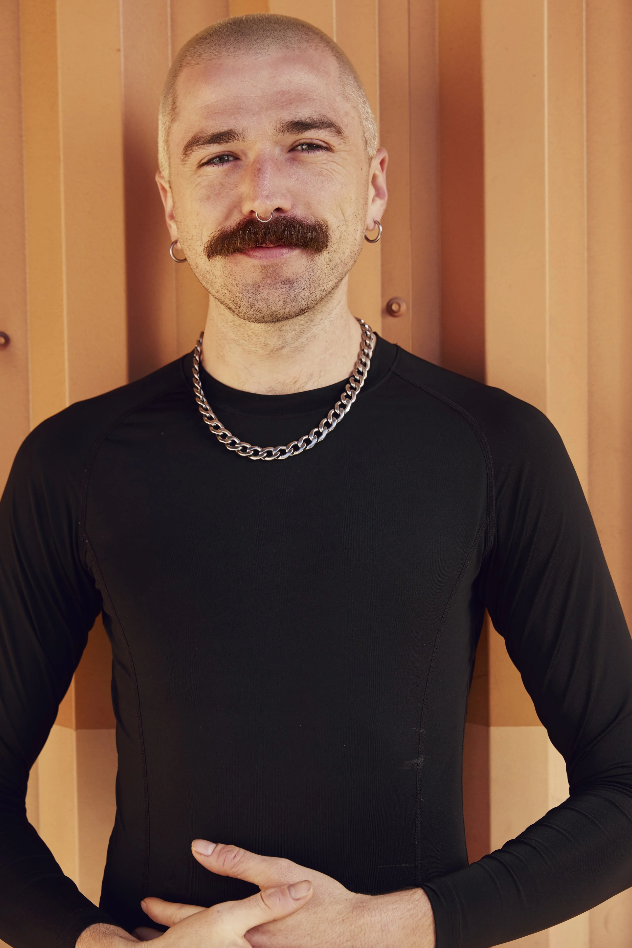 A man with a shaved head, gray eyes, a mustache, and a septum piercing, wearing earrings, a black leather jacket, and silver necklaces, against a plain neutral background.