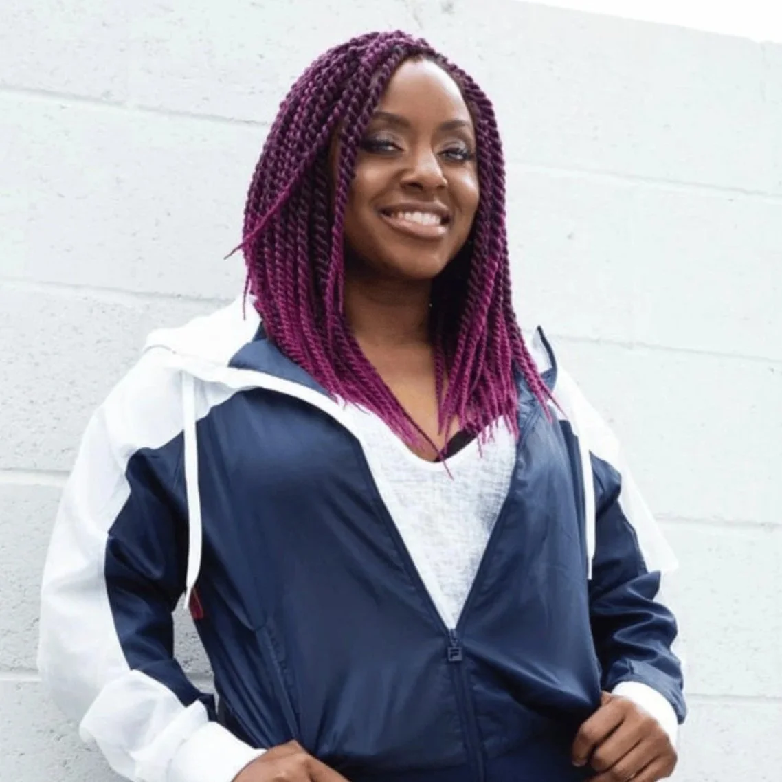A woman with dark skin, purple braided hair, smiling, wearing a white and navy blue jacket and standing in front of a white wall.