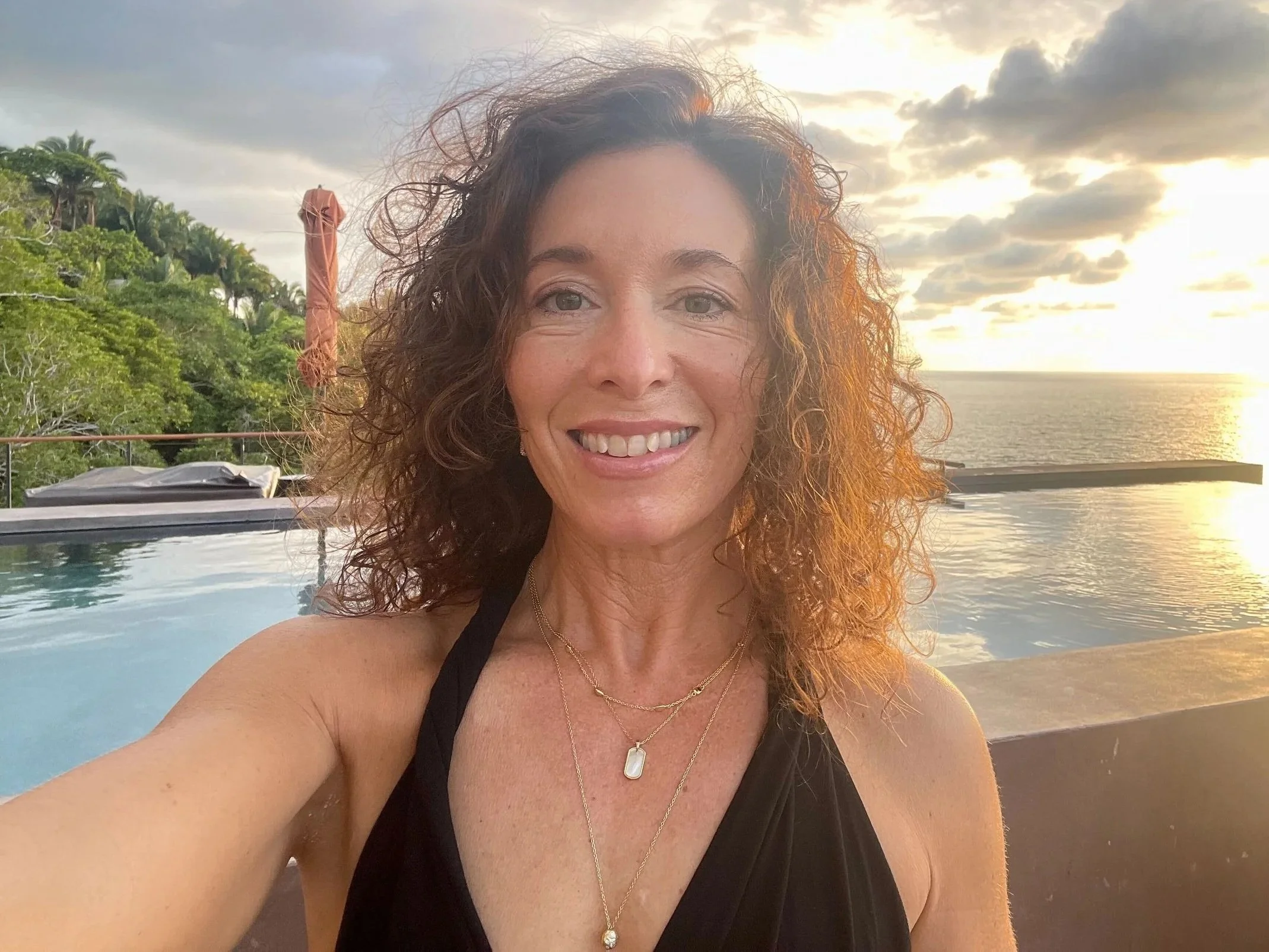 A woman practicing yoga outdoors, stretching with one arm raised behind her head and the other extended forward, smiling at the camera, with lush greenery in the background.