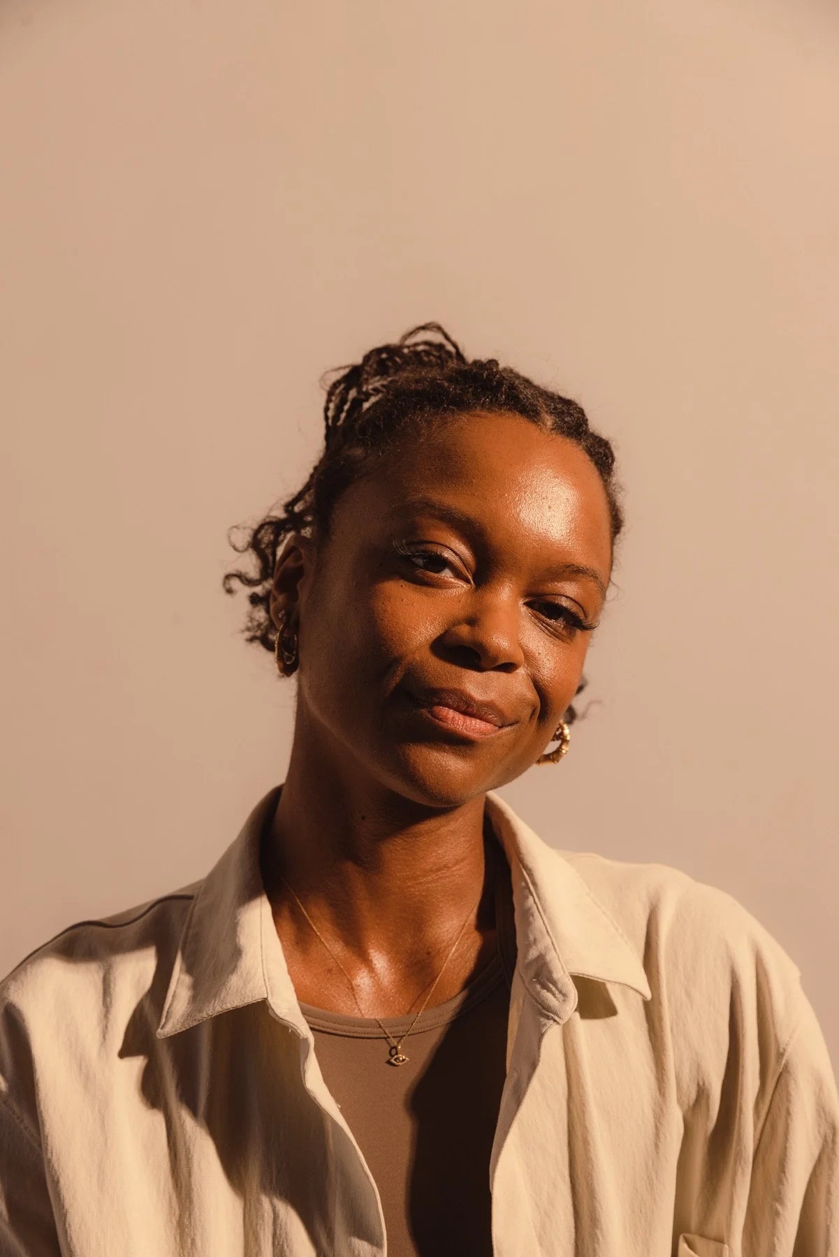 A woman with dreadlocks, wearing a black and white striped sweater, black pants, and black combat boots, sitting cross-legged against a plain white wall, smiling and holding her right hand to her chest.