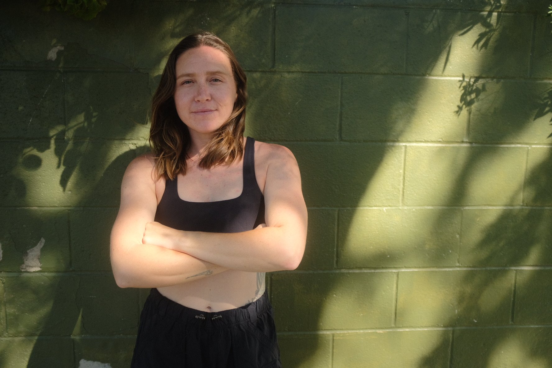 A woman with shoulder-length brown hair standing against a green painted brick wall, arms crossed, looking at the camera, with sunlight and shadows cast on the wall.