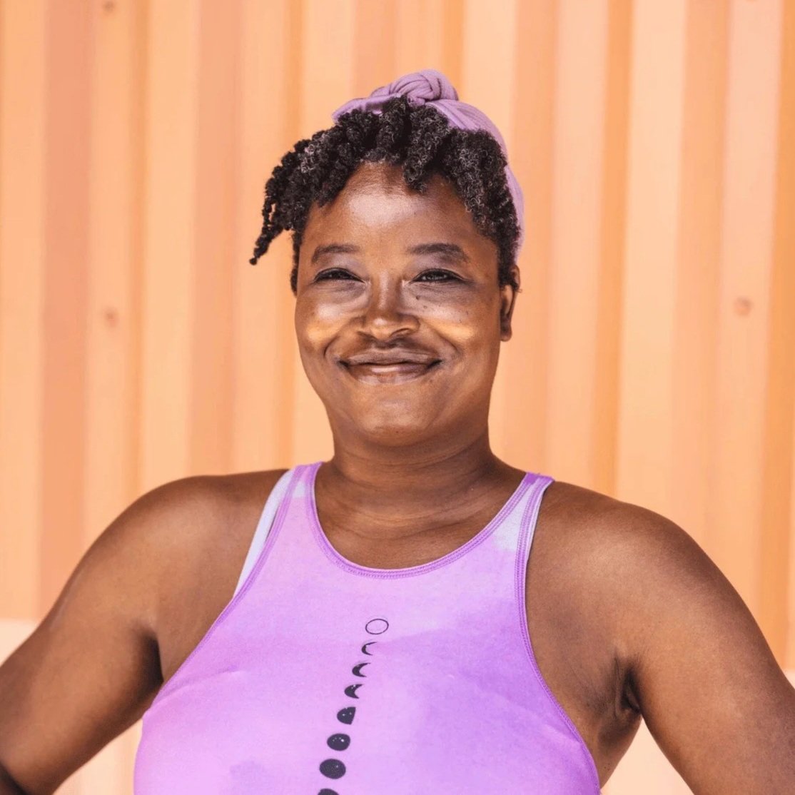 A woman smiling, wearing a purple sports top and headband, with a wooden background.