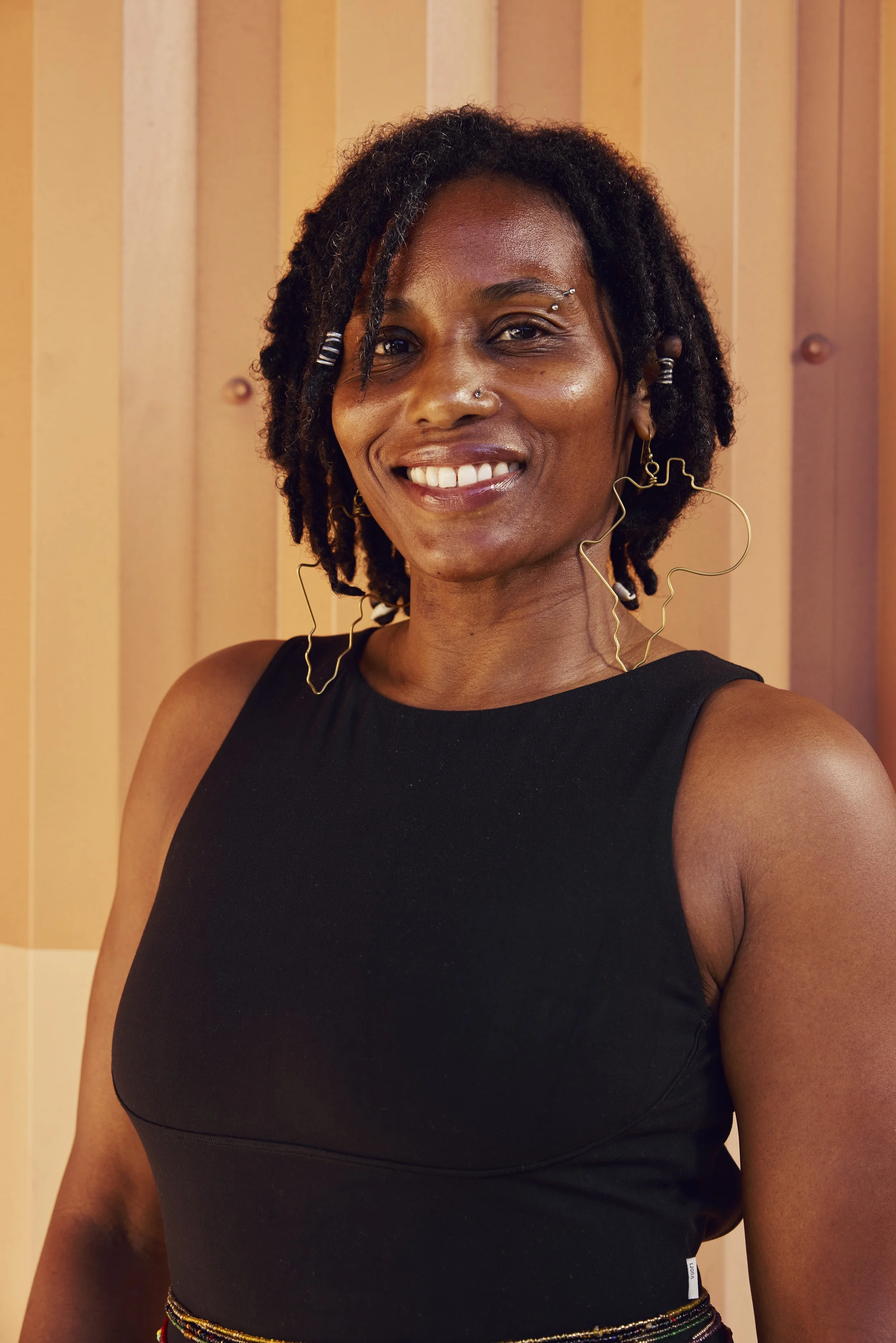 A woman with short, curly hair smiling, with her hand resting under her chin, wearing a patterned top and a smartwatch on her left wrist, in an indoor setting.