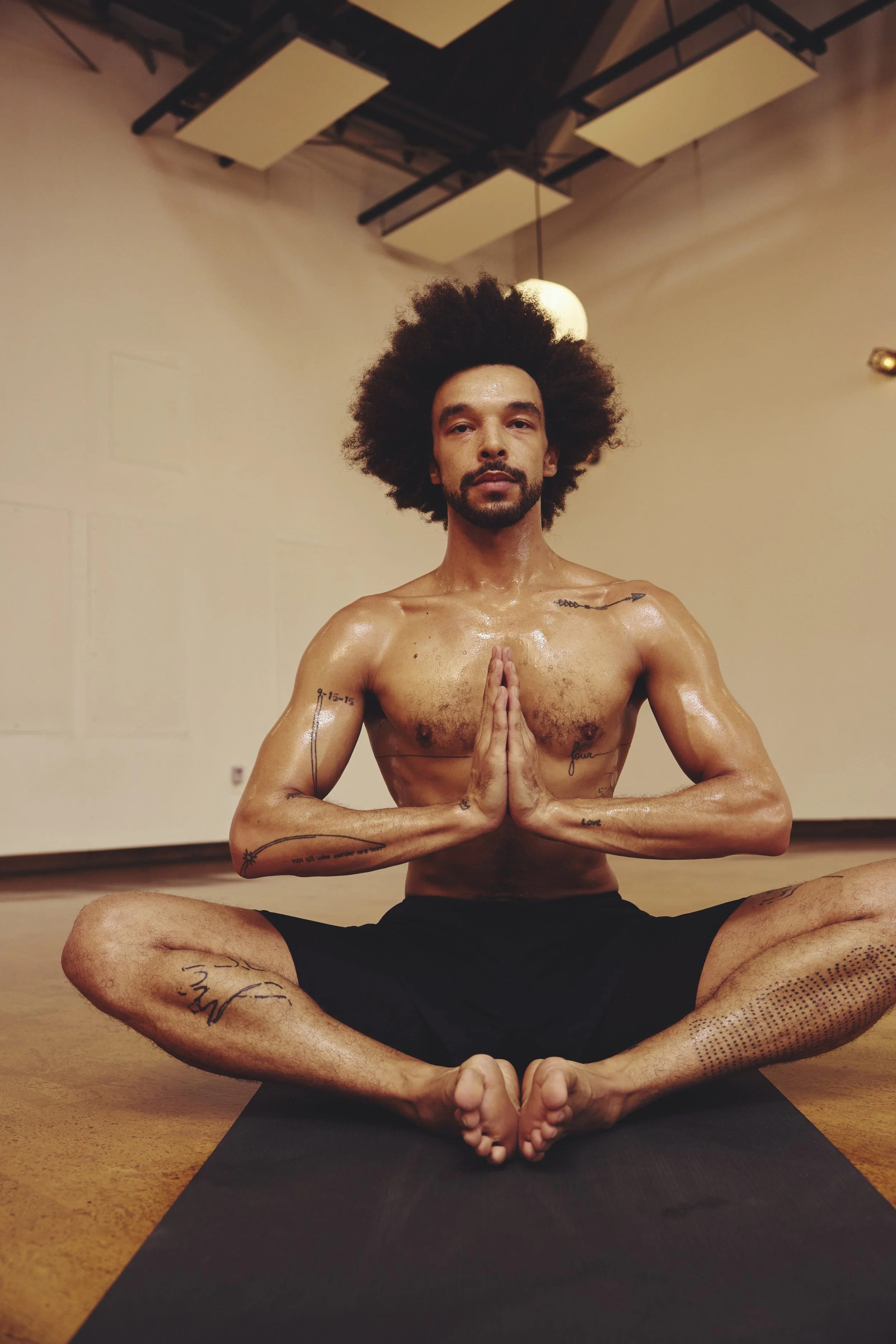 A shirtless man with tattoos sitting cross-legged on a yoga mat in a yoga studio, with hands in a prayer position and an afro hairstyle.