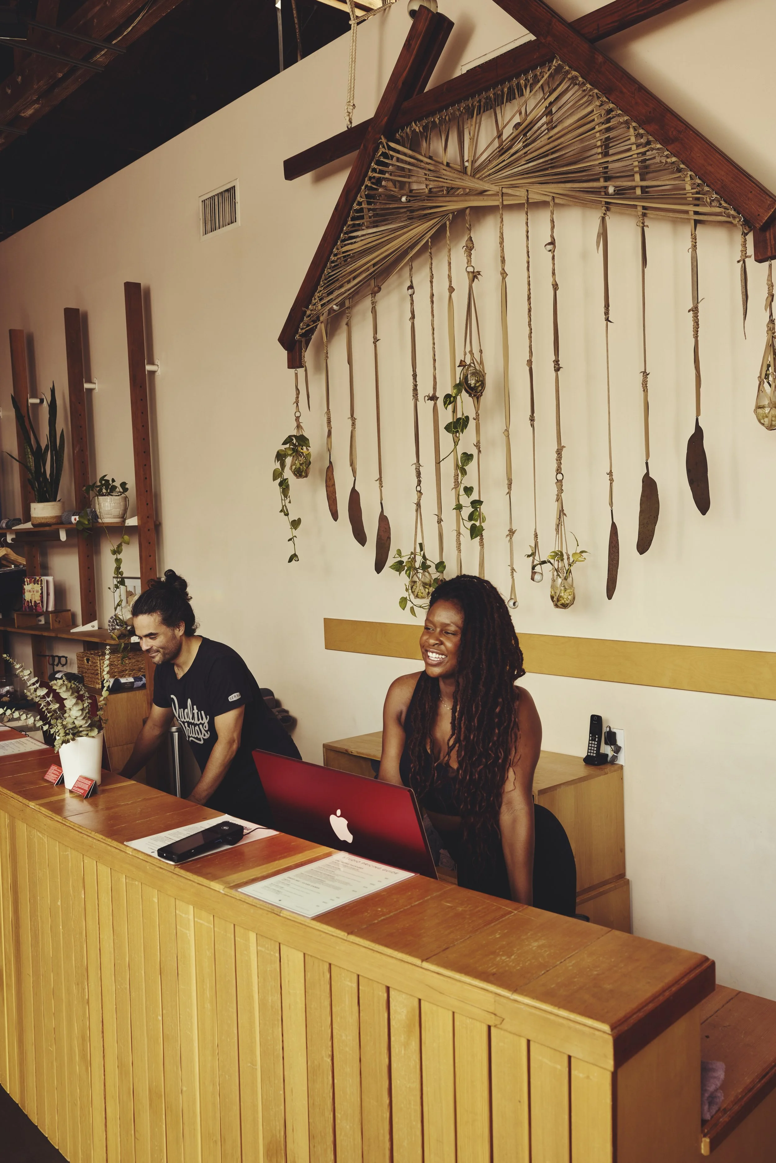 Two people sitting behind a wooden reception desk, a man and a woman, in a cozy, bohemian-style interior with wall art made of hanging rope, shells, and plants.
