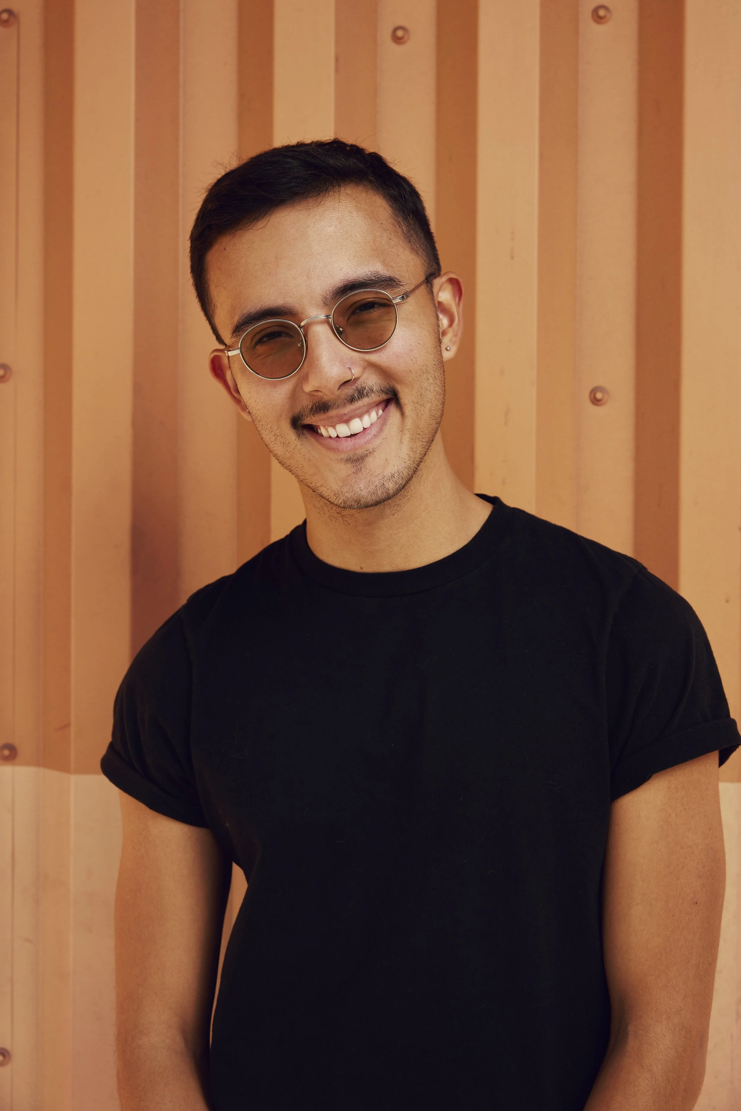 A young man with short dark hair, wearing round sunglasses, a septum piercing, earrings, and a black t-shirt, smiling in front of a wooden wall.
