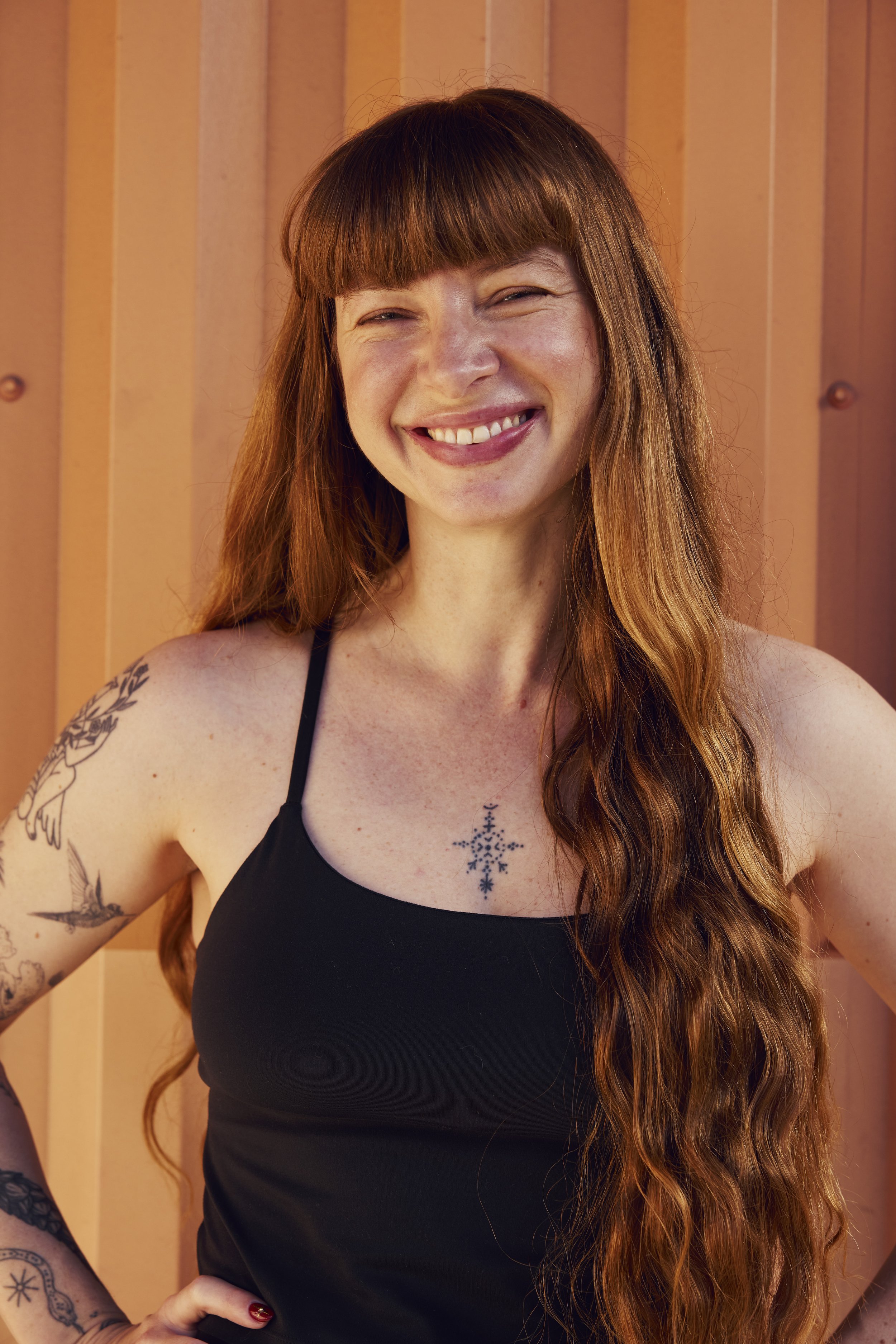 A woman with long wavy hair and tattoos on her shoulder stands outdoors in a desert landscape with Joshua trees, smiling at the camera during sunset.