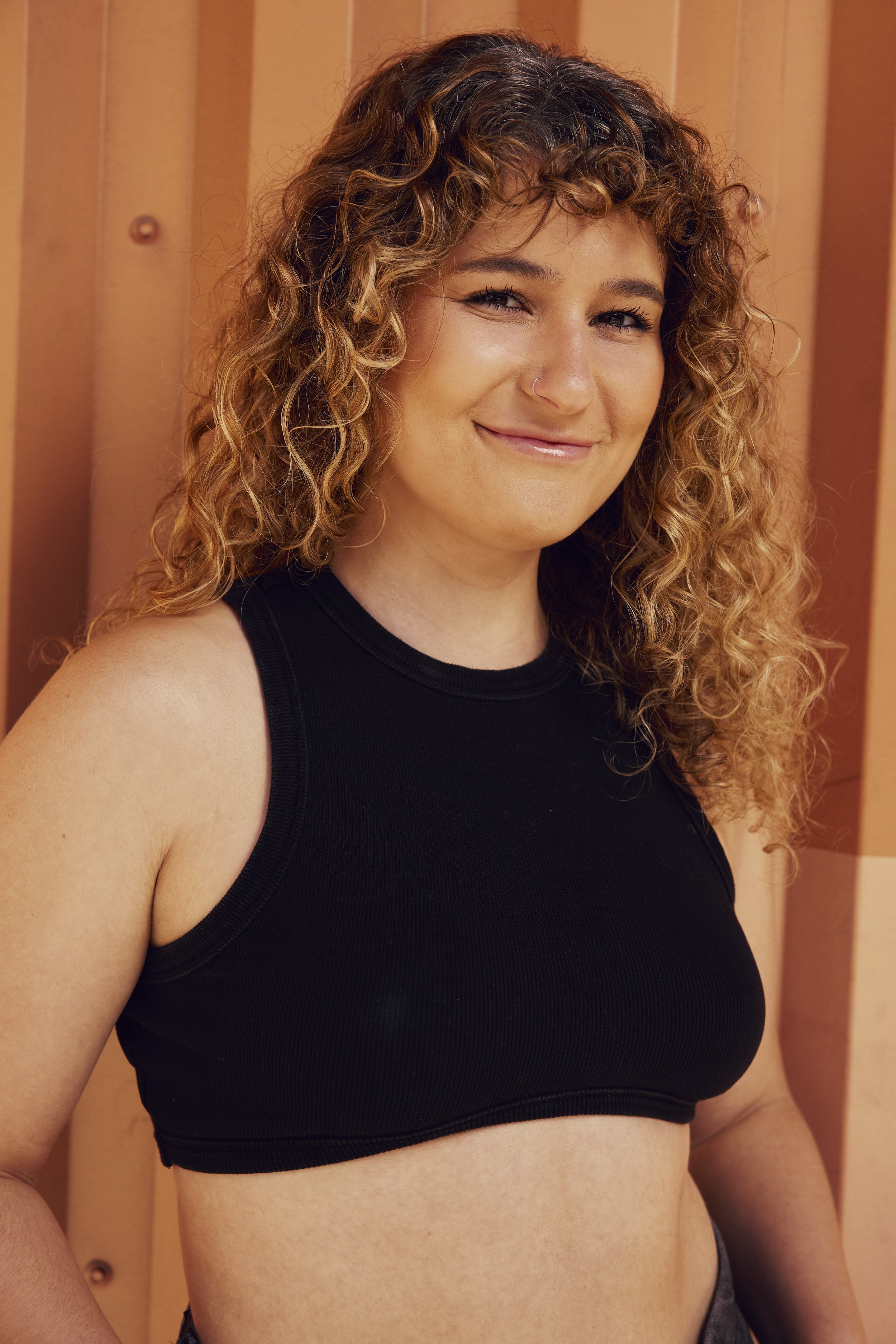 Portrait of a young woman with curly hair smiling, wearing a blue swimsuit, against a plain white background.