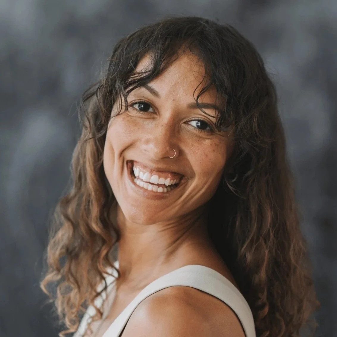 A woman smiling with curly brown hair, freckles, and a nose ring, looking at the camera against a blurred dark background.