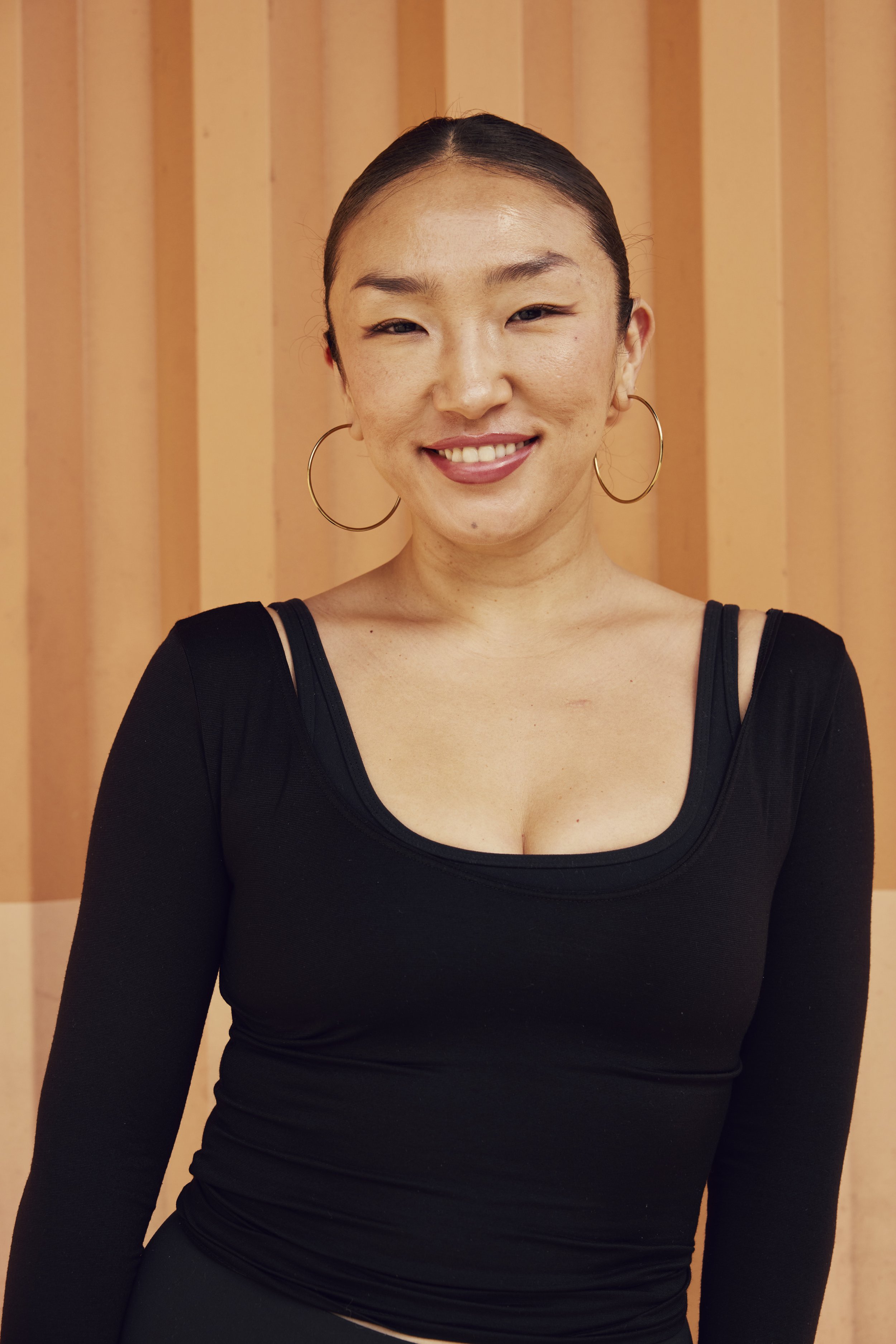 A woman with dark wavy hair, wearing hoop earrings, a septum ring, and a dark gray spaghetti strap top, smiling and laughing against a purple background.