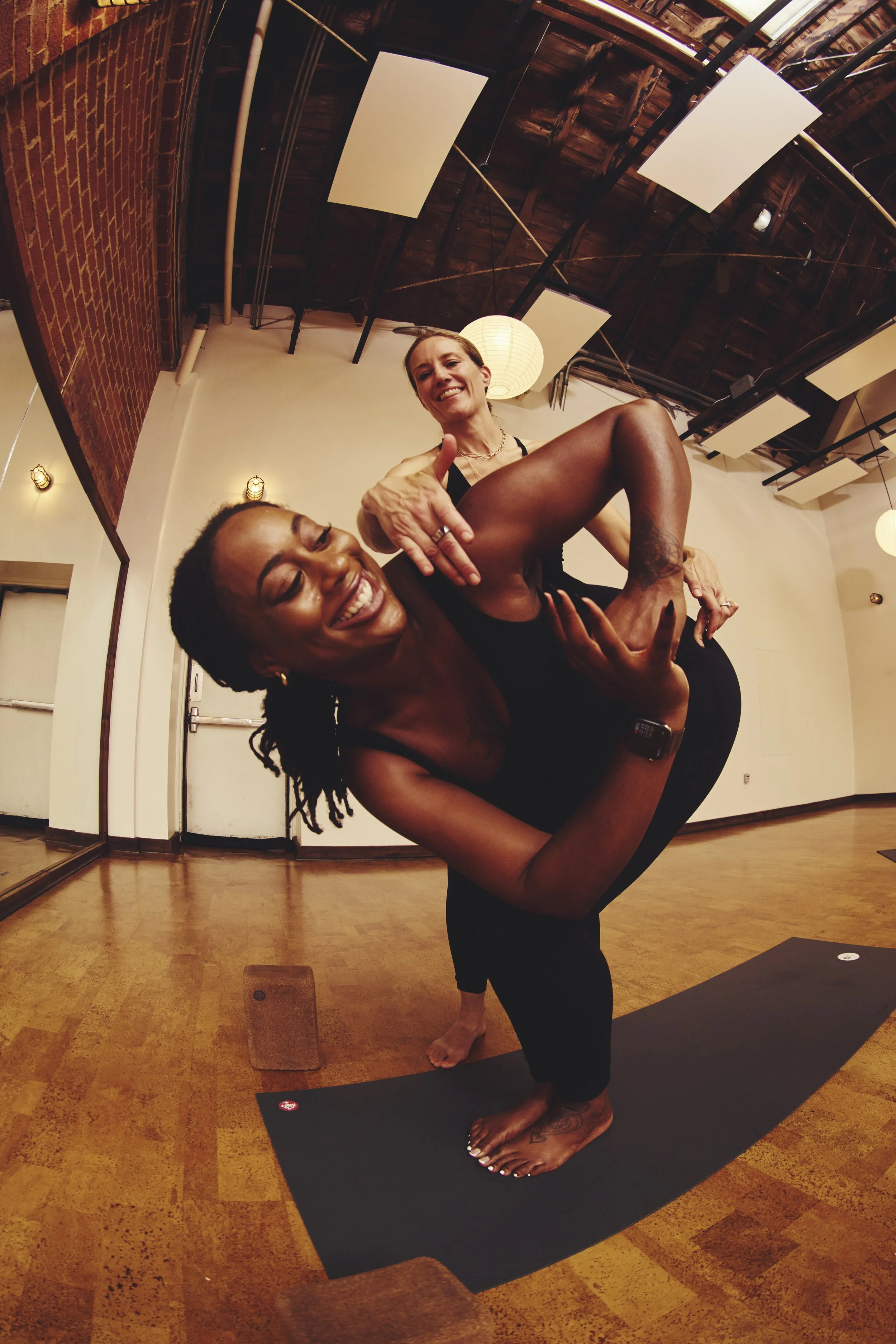Two women practicing yoga in a studio, one in a yoga pose holding the other in a balanced pose, both smiling.