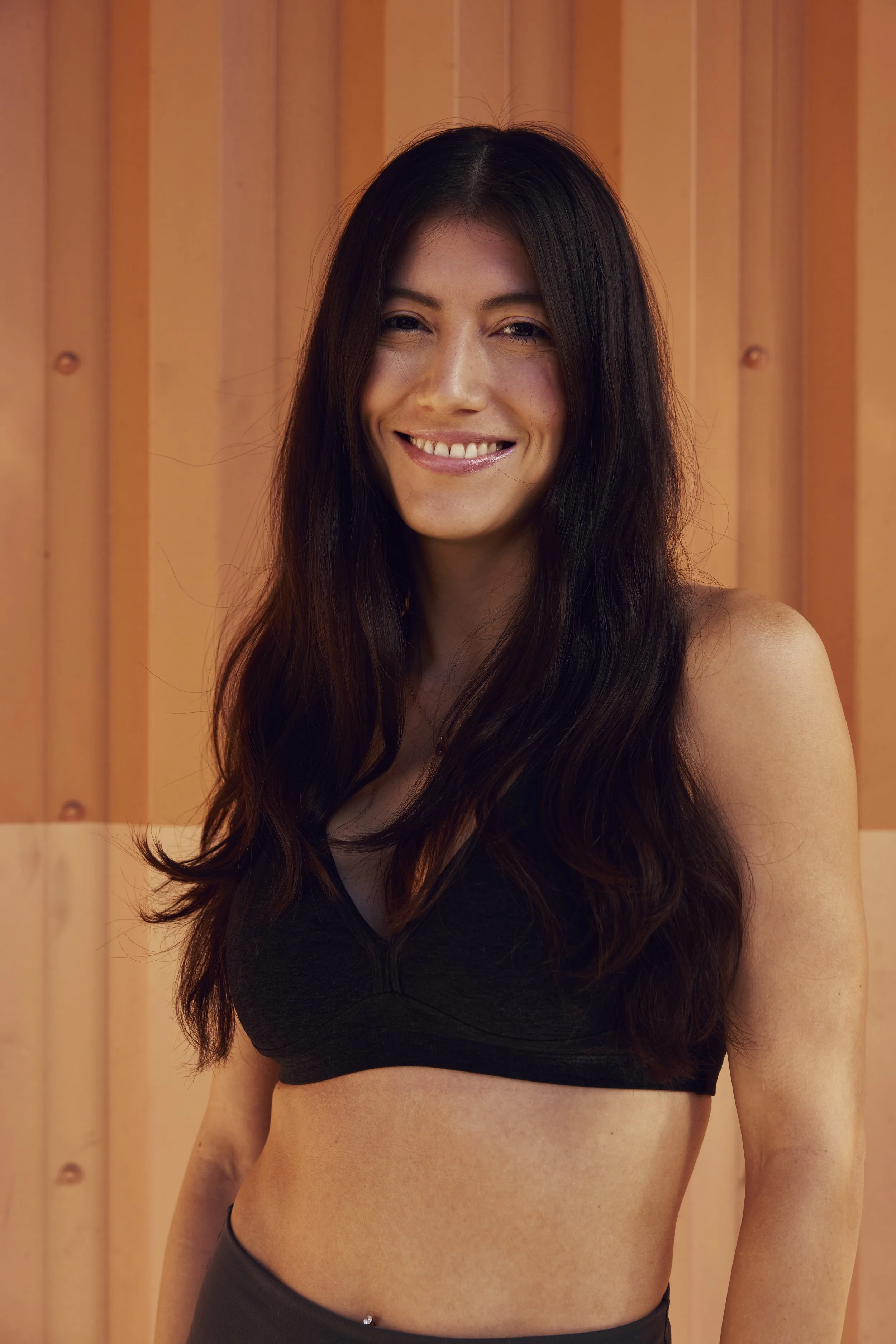 A woman with long dark hair wearing hair clips, smiling on a beach with ocean waves in the background.
