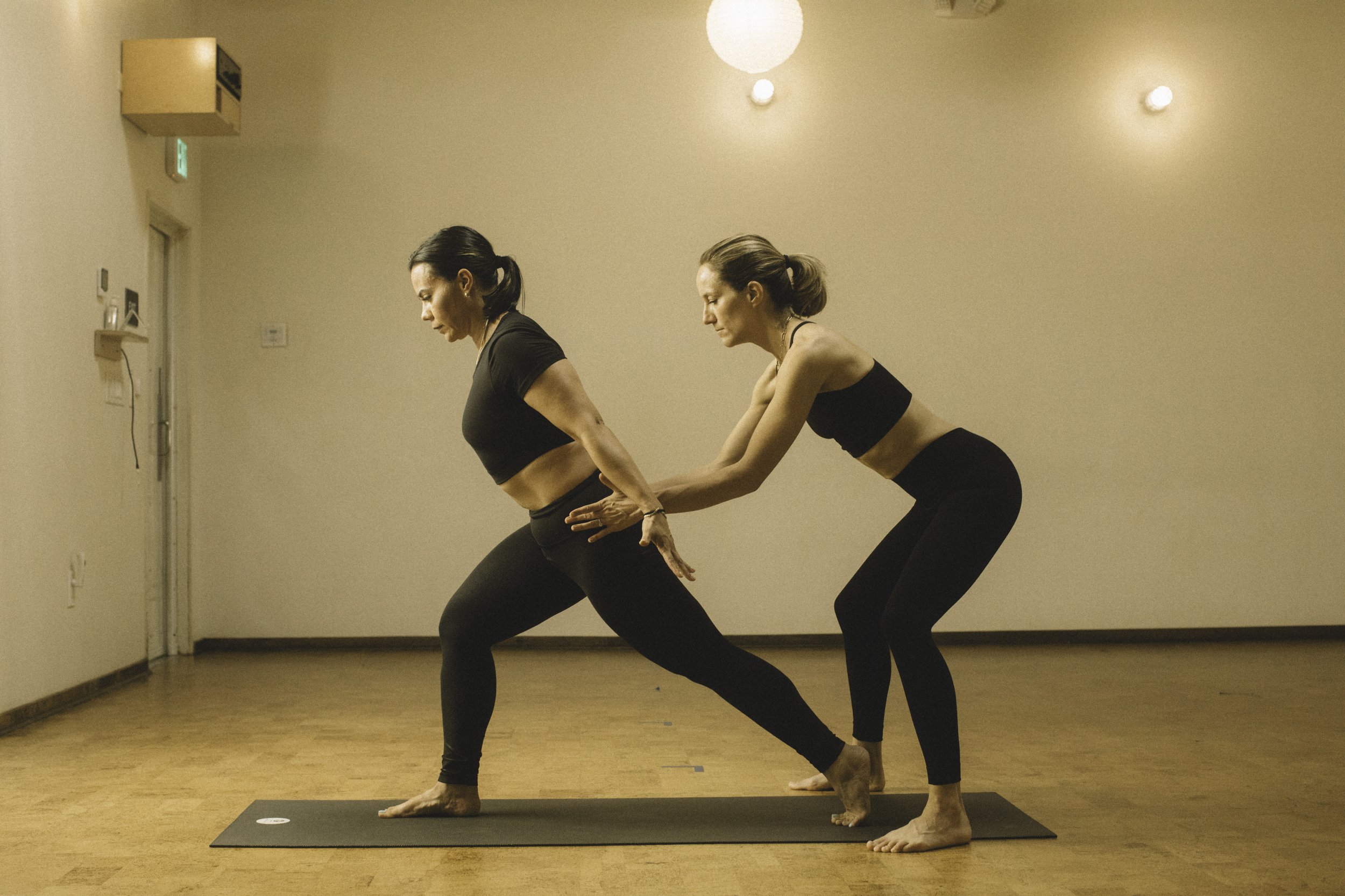 Two women practicing yoga together in a studio. One woman is in a squat position, and the other woman is supporting her from behind.