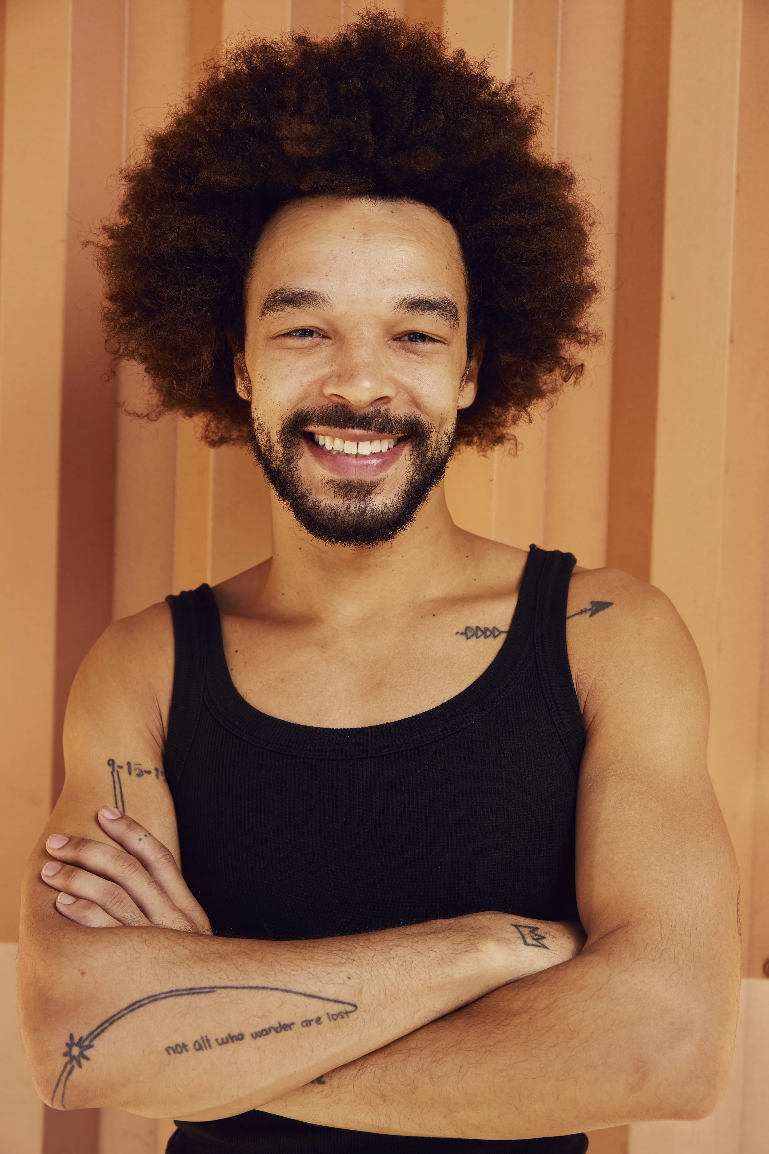A smiling man with a big, curly hairstyle, a beard, and a mustache, wearing a dark t-shirt, standing against a plain white wall.