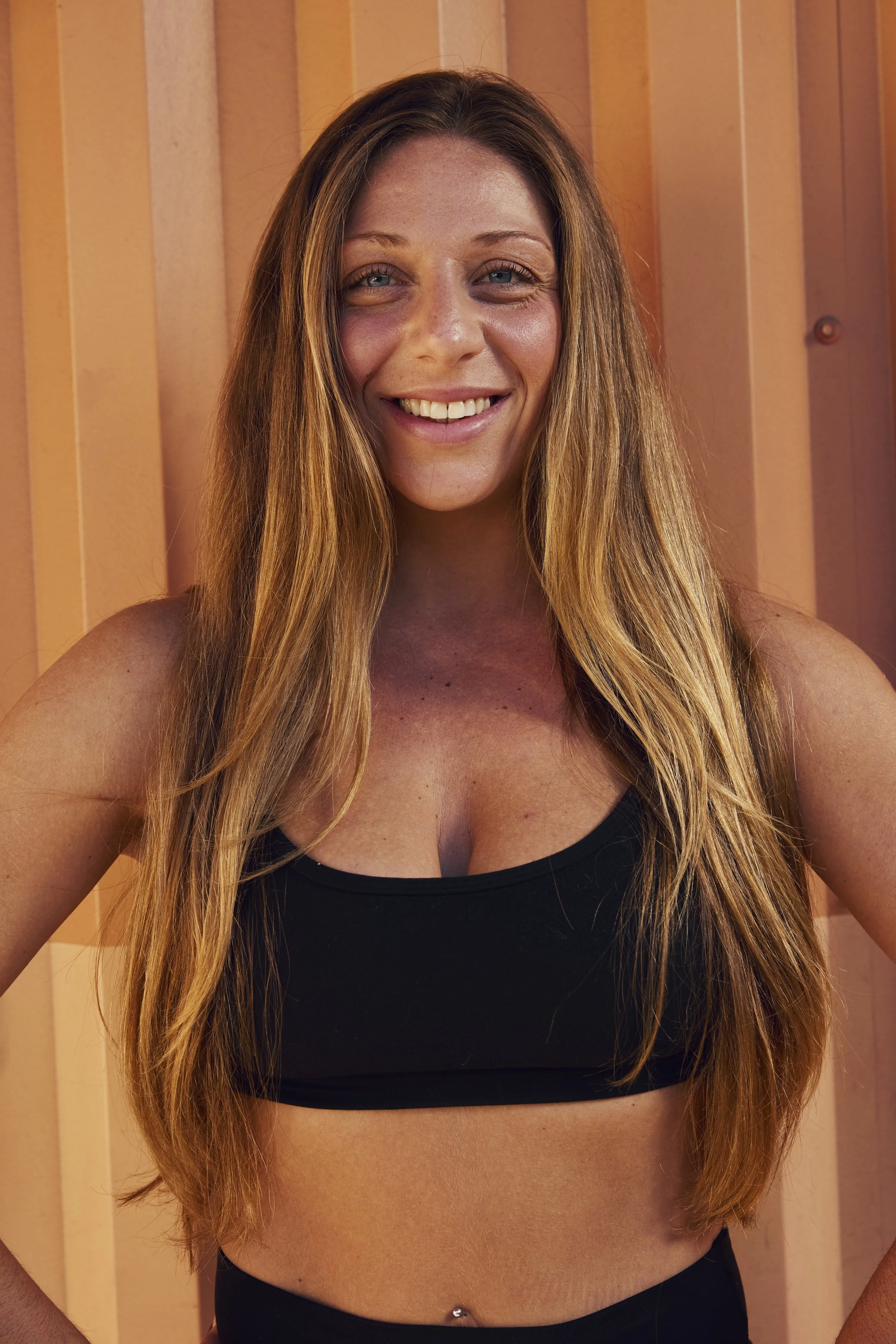 Close-up of a woman with long wavy brown hair and blue eyes, smiling at the camera in a restaurant or cafe setting.