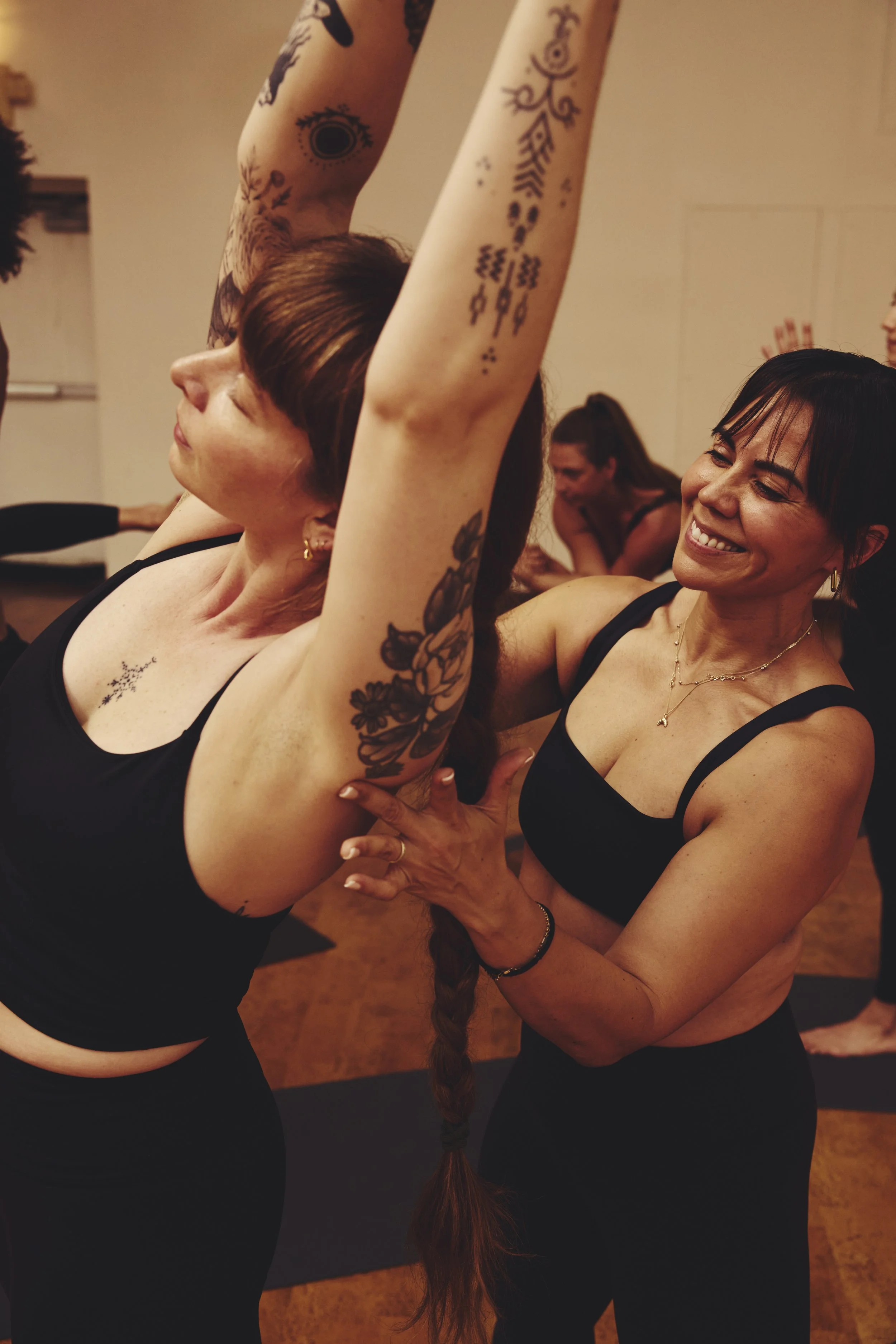 Two women practicing acroyoga indoors, with one lifting the other, who has her arms extended overhead. Both women are smiling and wearing black tops, with tattoos visible on the woman being lifted.