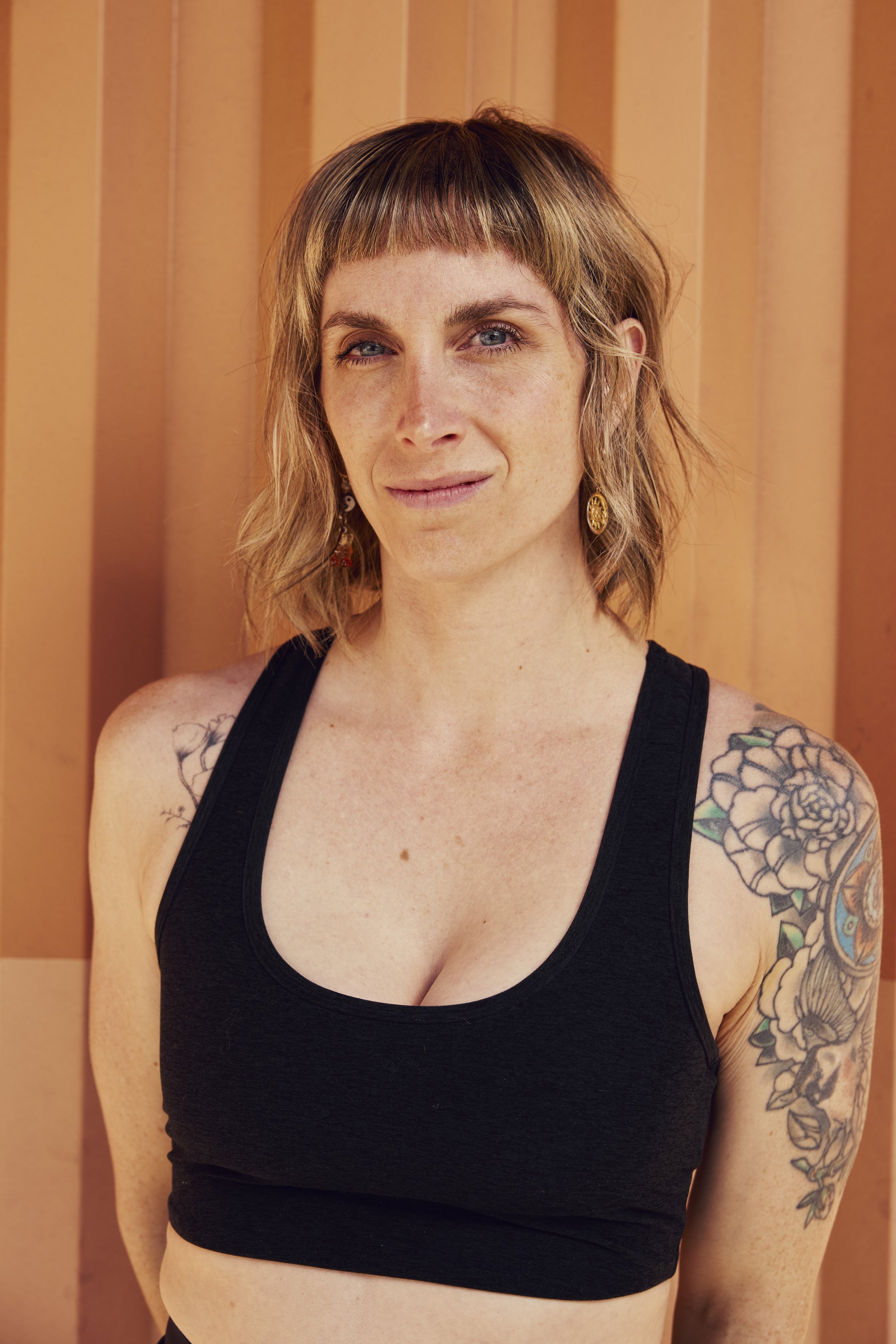 Black and white photo of a woman with dark hair in a bun, wearing earrings and a patterned blouse, standing with her arms crossed in front of a metallic background.
