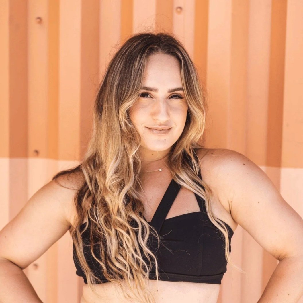 A woman with long, wavy blonde hair wearing a black top posing against a wooden background.
