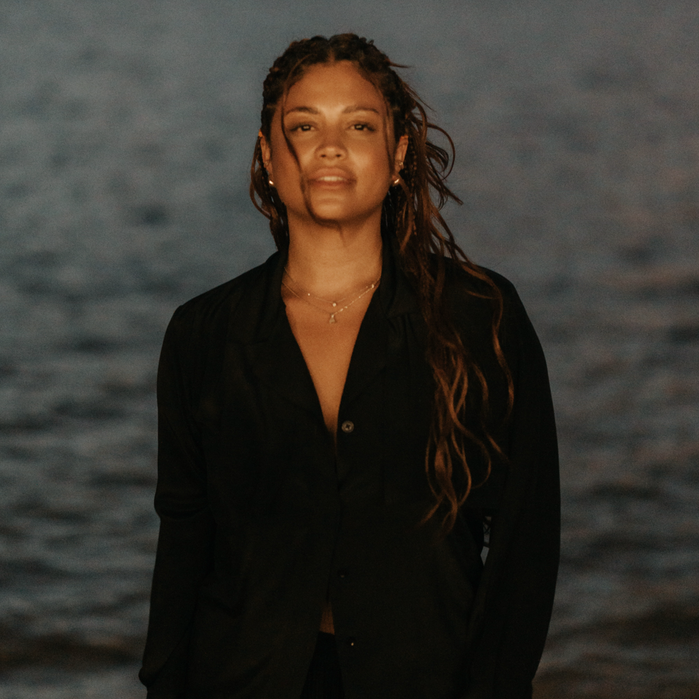 A woman with long black hair, eyes closed, and right hand on chest, sitting in front of a large metallic circular object, under warm yellow-orange lighting.