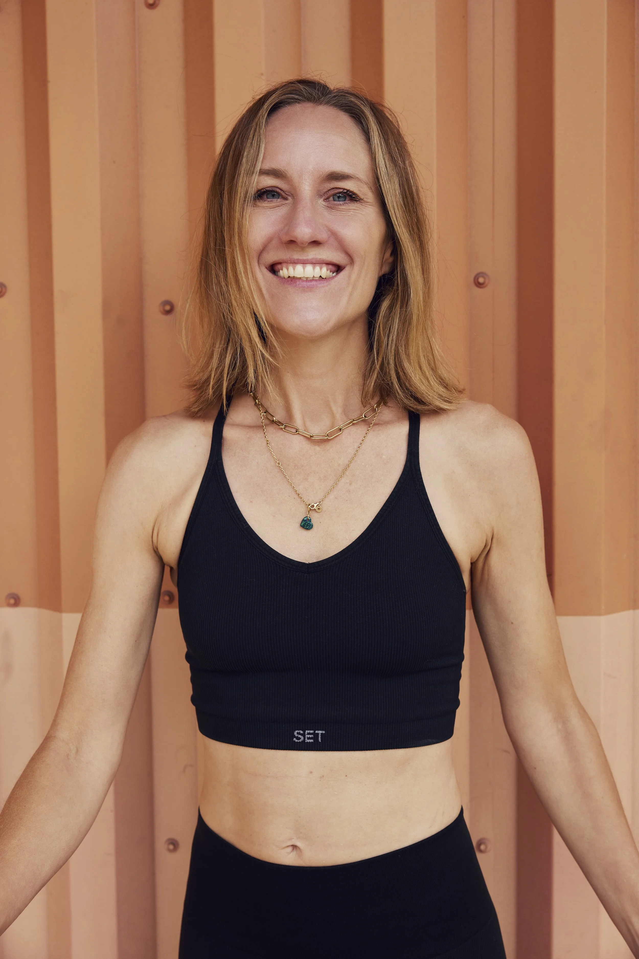 A woman with shoulder-length blonde hair, smiling, wearing an orange sleeveless top, standing in front of a peach-colored wall with vertical panels.