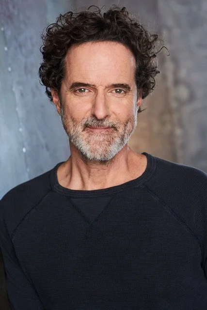 A middle-aged man with curly dark hair and a beard, wearing a black sweater, standing in front of a textured gray and blue background.