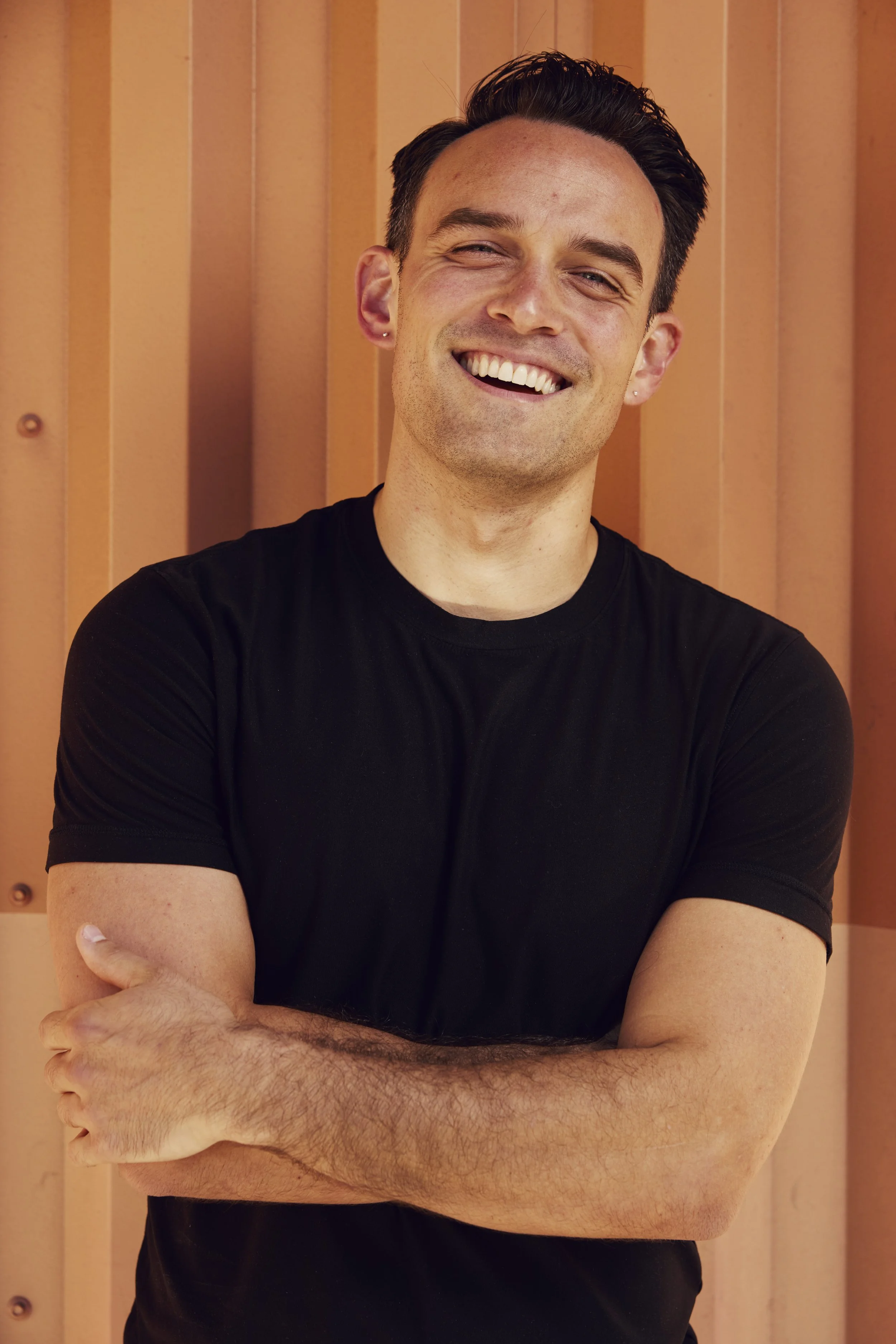 A young man with short dark hair, blue eyes, and light skin, wearing a black v-neck shirt, stands in front of a wooden background.
