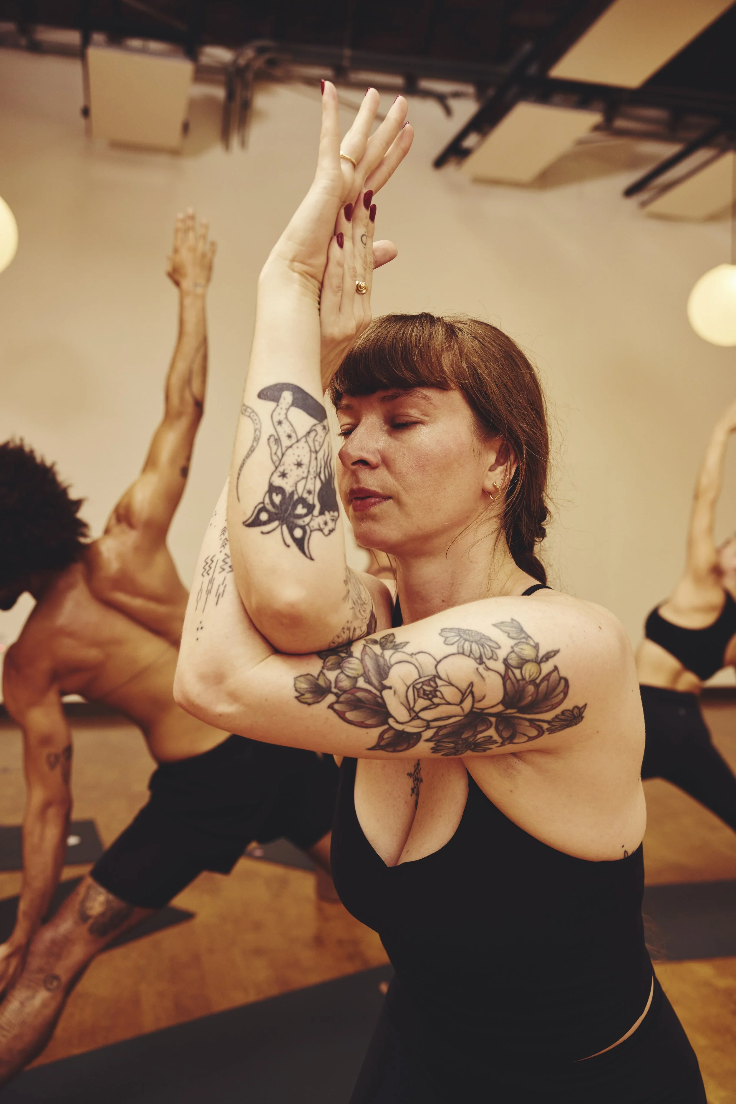 Women practicing yoga in a studio, wearing athletic clothing, with tattoos on their arms, and focusing on a pose with closed eyes.