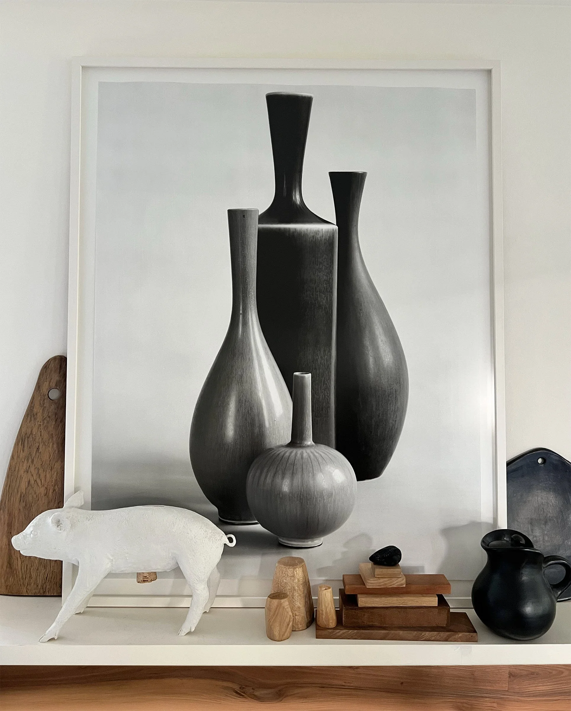 Decorative tabletop arrangement with framed black and white photo of vases, white pig sculpture, wooden blocks, small black stones, and black pitcher on a white surface.