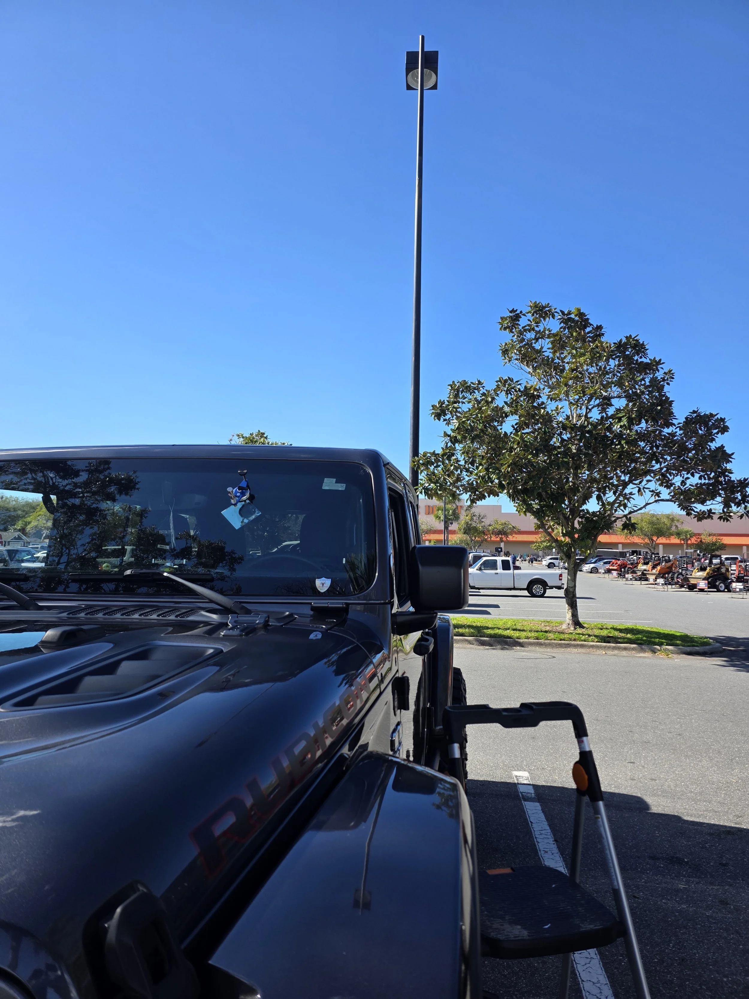 Black Jeep parked in a parking lot next to a small step ladder, with a tree and other parked vehicles in the background under a clear blue sky.