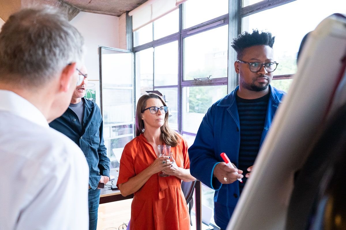 Sarah Bellorini, Leadership Identity Coach for Gen X, with three other people in a meeting room, with large windows, discussing around a large whiteboard.