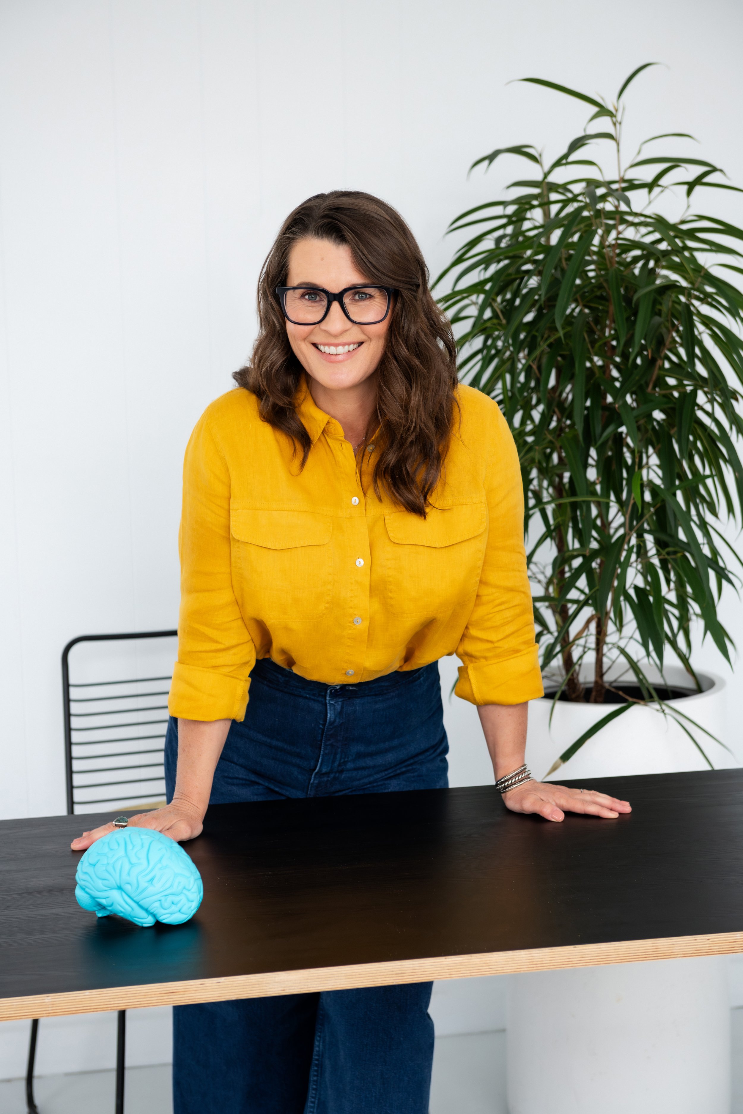 Sarah Bellorini, Leadership Identity Coach for Gen X, leaning on a table with her hands, smiling at the camera. There is a plant and a chair behind her, and a blue brain model on the table.