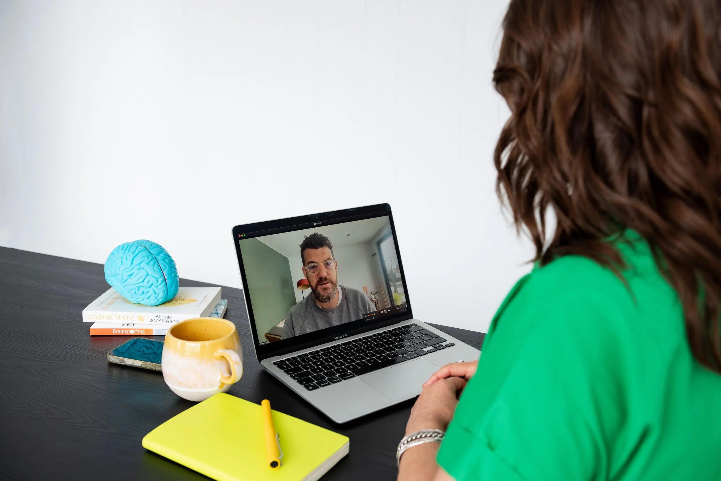 Sarah Bellorini, Leadership Identity Coach for Gen X, video chatting with a man on a MacBook laptop. On the desk there's a yellow and white mug, a smartphone, a blue brain-shaped object on top of books, and a framed picture of a brain.