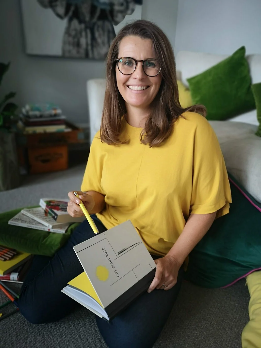 Sarah Bellorini, Leadership Identity Coach for Gen X, smiling at the camera, surrounded by books and green cushions in a living room.