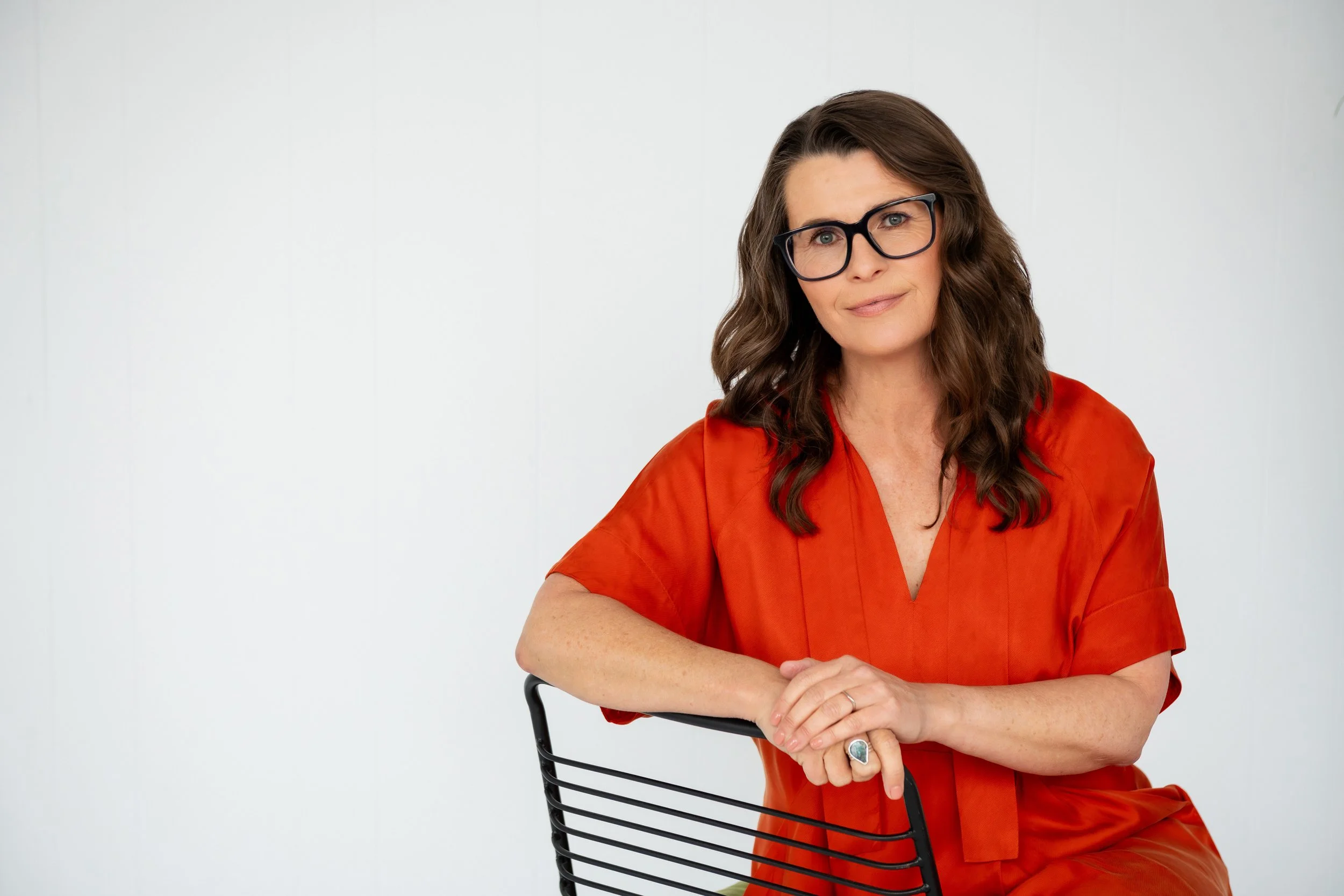 Sarah Bellorini, Leadership Identity Coach for Gen X, wearing an orange top, sits on a black chair with a white background.