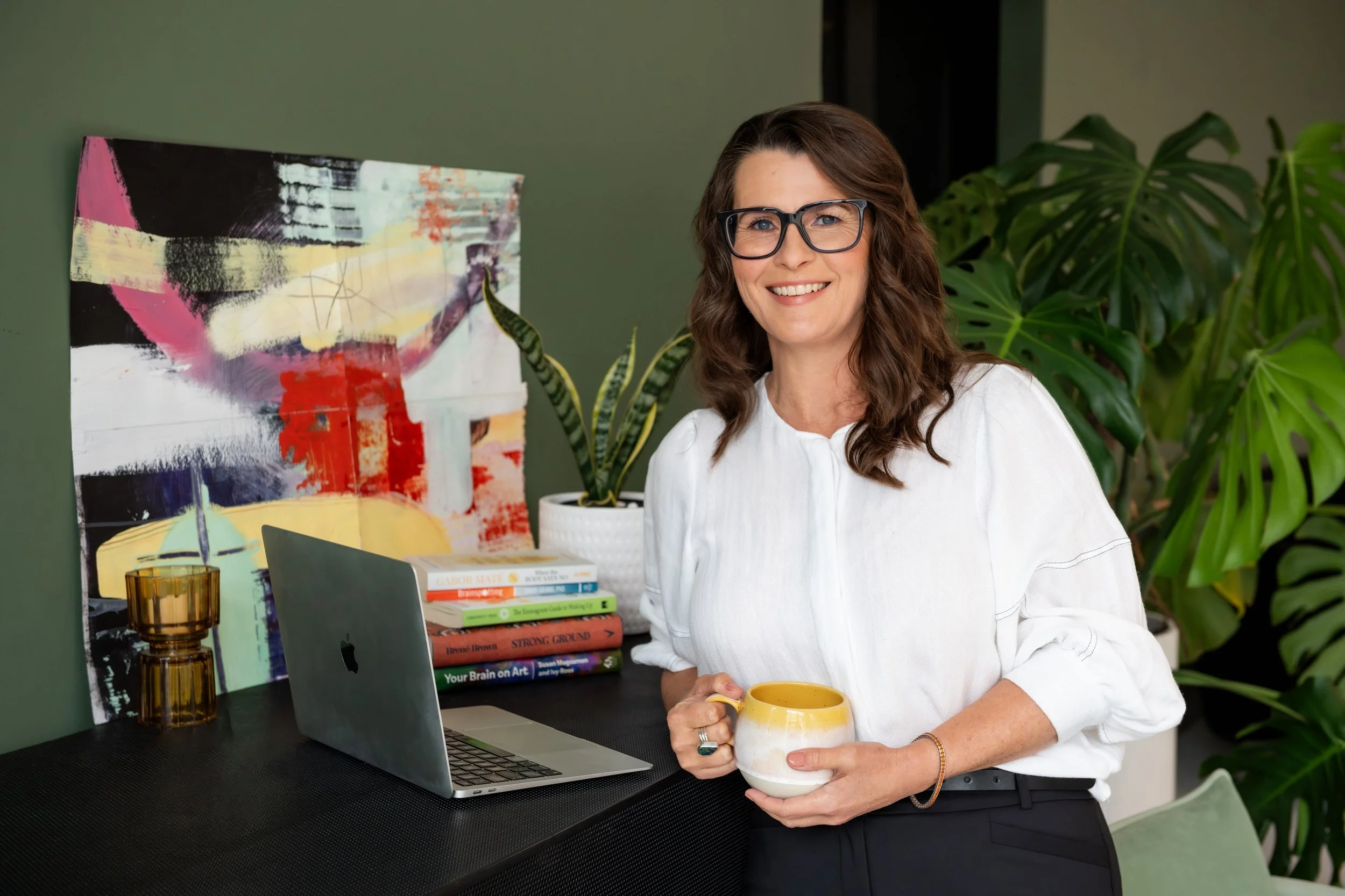 Sarah Bellorini, Leadership Identity Coach for Gen X, smiles while holding a yellow mug, standing in front of a desk with books, a laptop, and a painting, surrounded by houseplants.