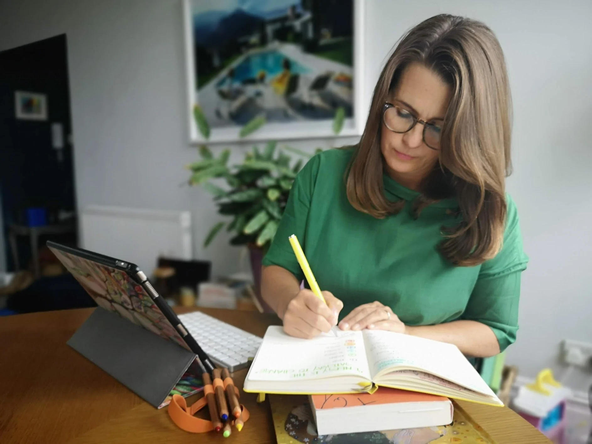 Sarah Bellorini, Leadership Identity Coach for Gen X, writing in a notebook with a yellow pen. There are colored pencils, a laptop, and stacked books on the table. A large potted plant is in the background, along with a piece of artwork and a shelf.
