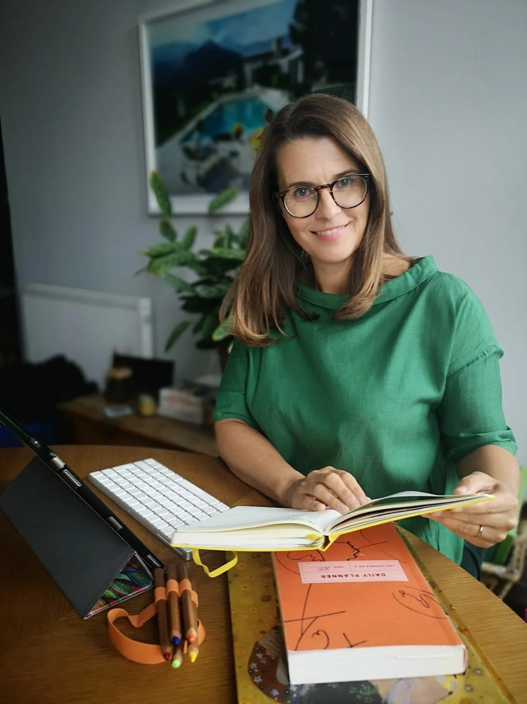 Sarah Bellorini, Leadership Identity Coach for Gen X, reads a book at a wooden desk. There is a keyboard, colored pens, and a folder on the desk, with a large houseplant and painting in the background.