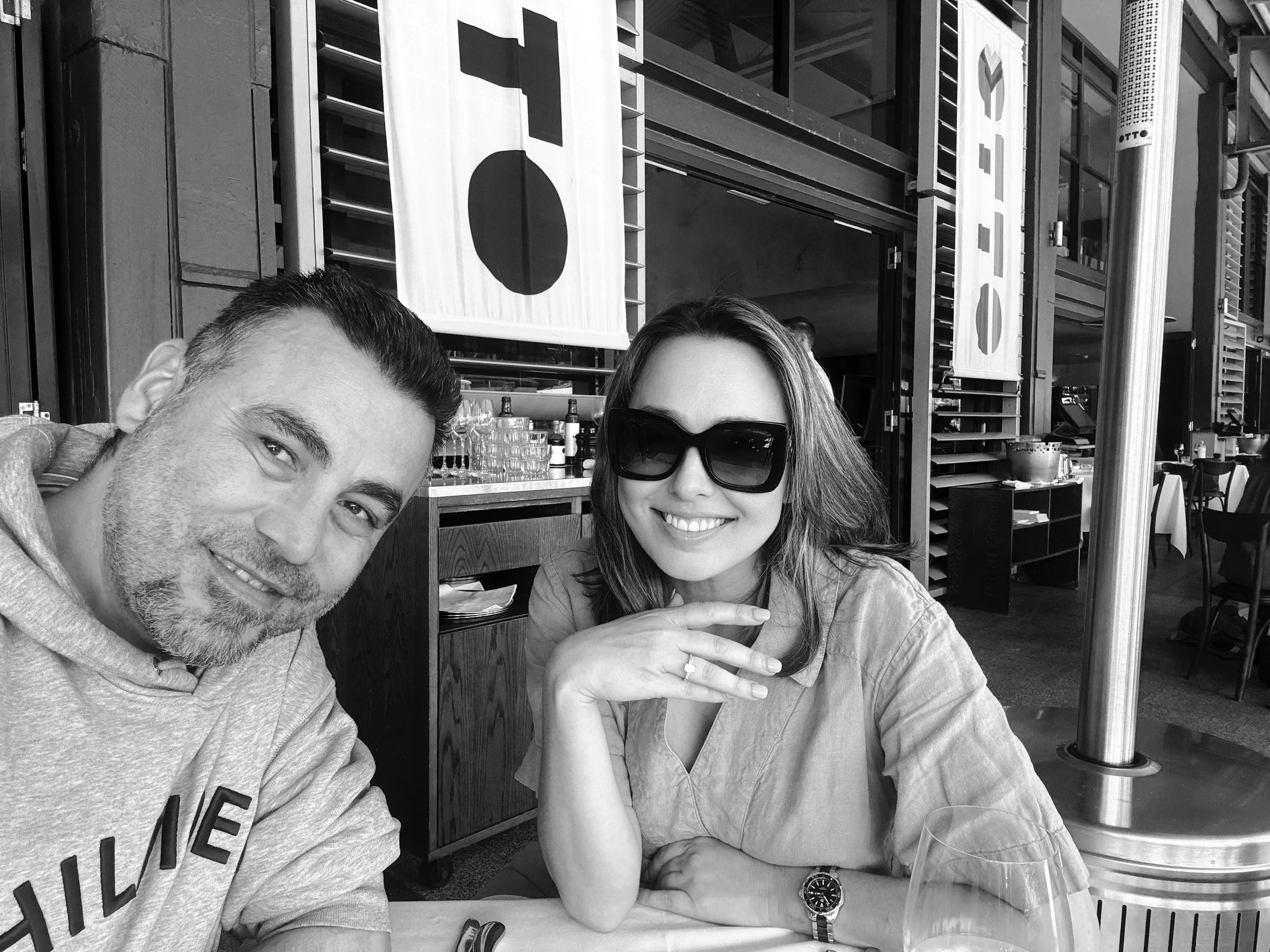 A black and white photo of a man and a woman sitting at a restaurant table. The man has short hair and a beard, wearing a hoodie, and is smiling at the camera. The woman has shoulder-length hair, large sunglasses, and a watch on her wrist, smiling with her hand resting under her chin.