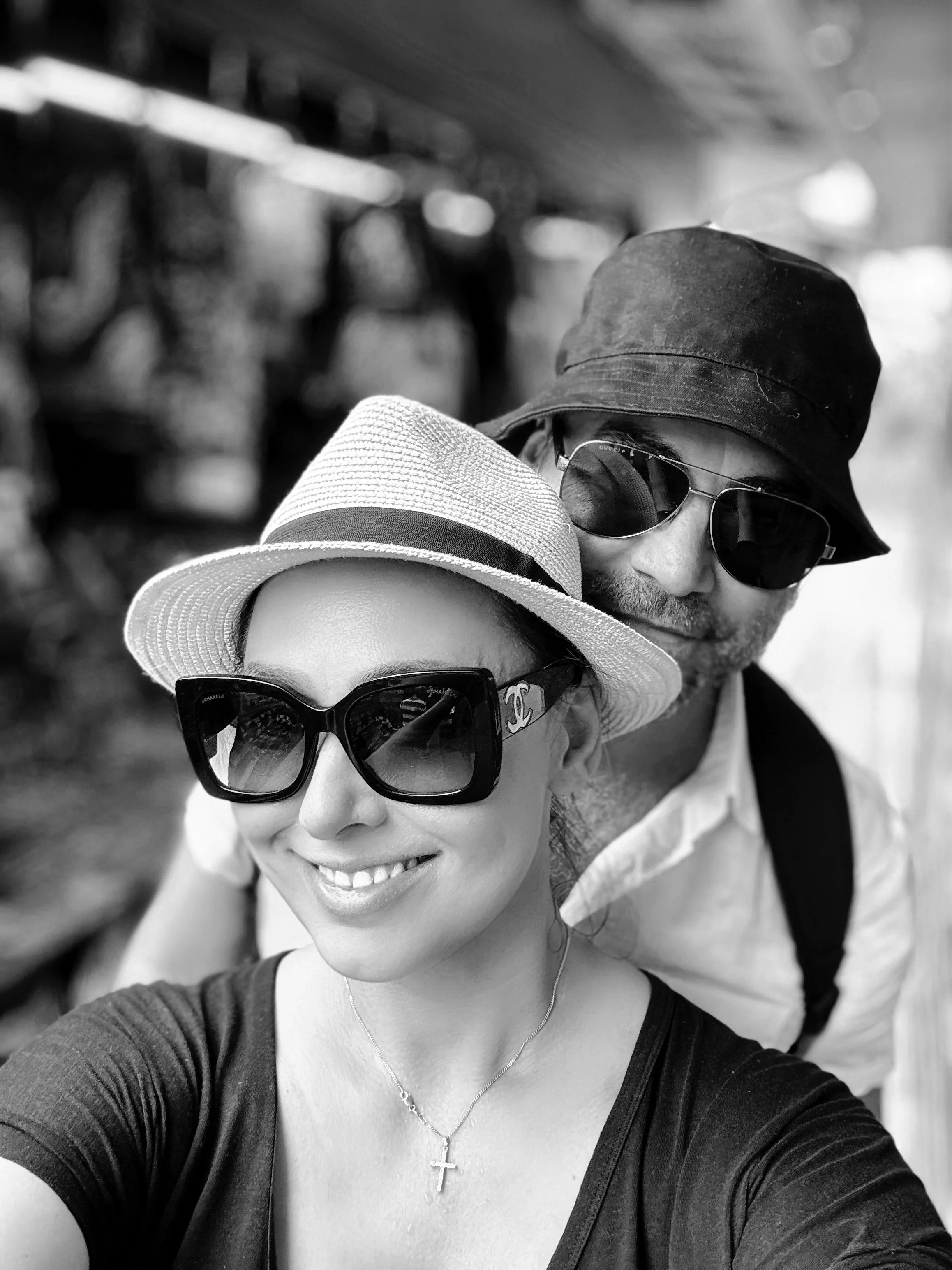 A woman and a man taking a selfie on a sunny day wearing sunglasses and hats, smiling, with a blurred background.