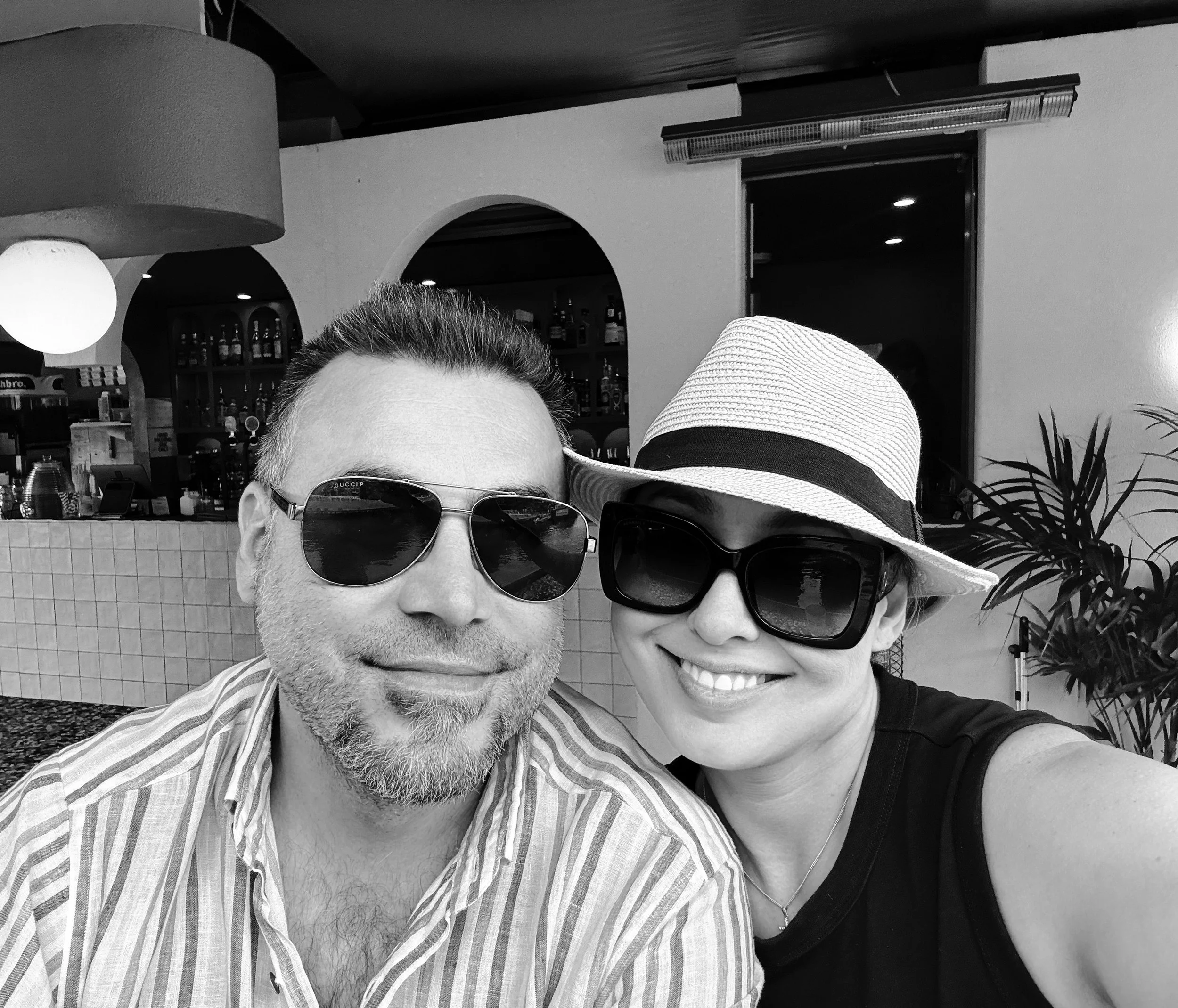 A man and woman taking a selfie together at a bar or restaurant. Both are wearing sunglasses and smiling, with the woman wearing a sunhat and a sleeveless top.
