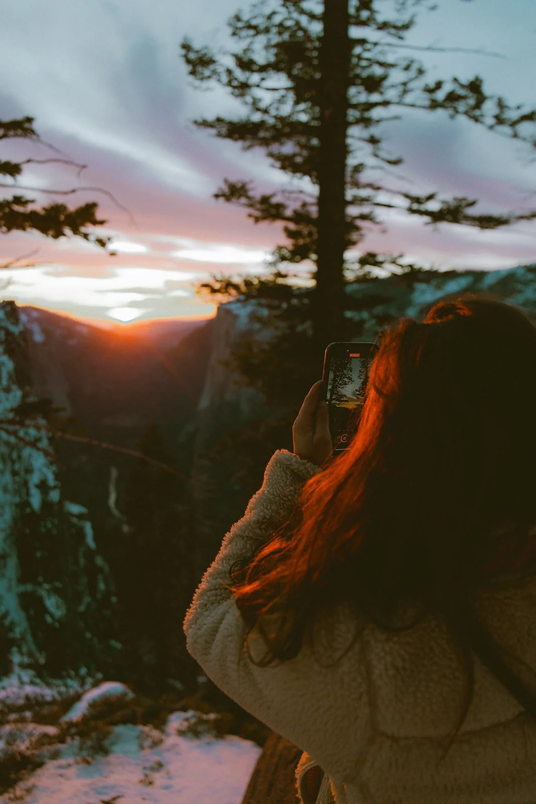 A woman with long hair taking a photo of the sunset over a canyon at a forested overlook during winter, with snow on the ground.