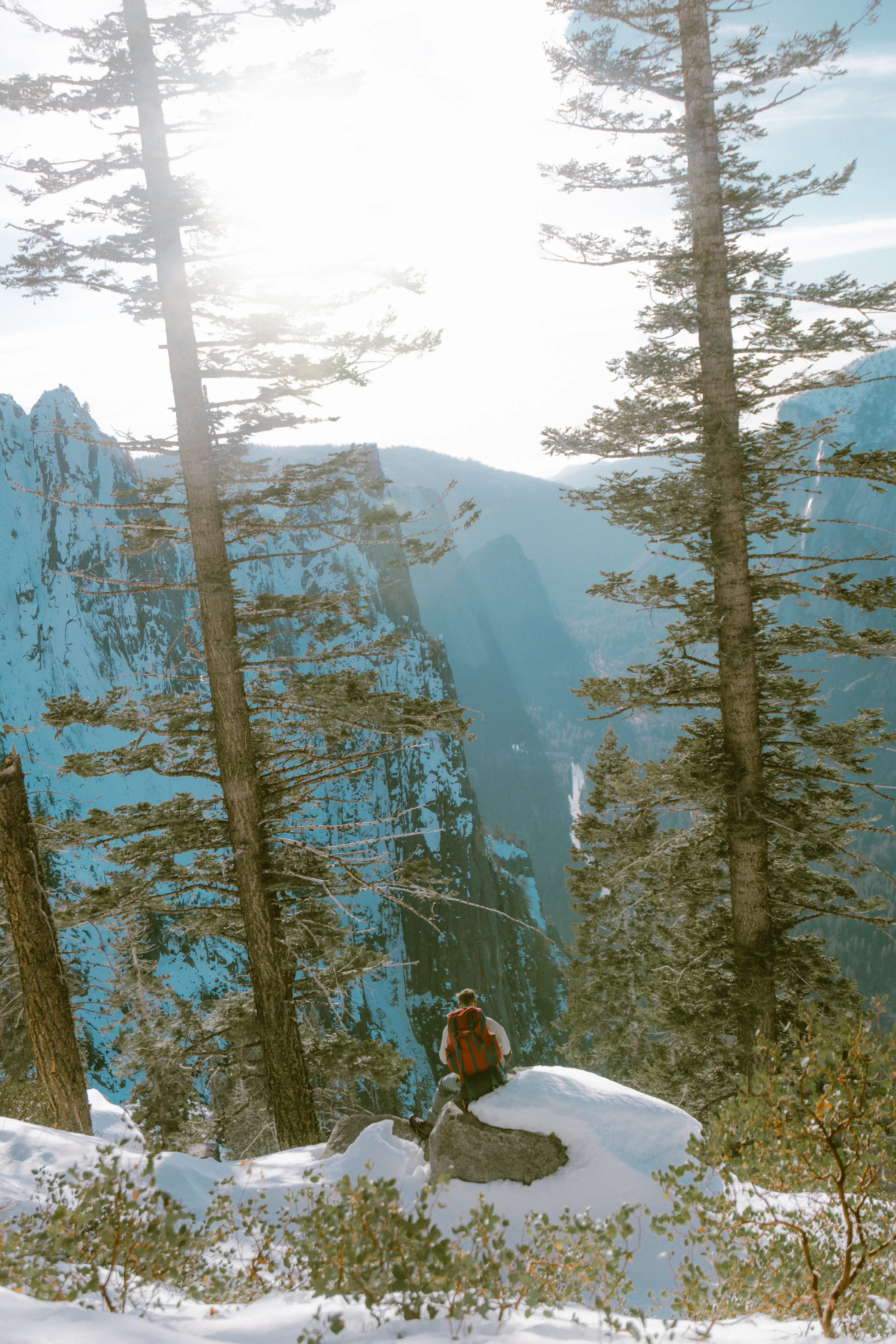 Hiker with a red backpack sitting on a rock in a snowy forest, overlooking a mountainous landscape with snow-covered peaks and tall pine trees.