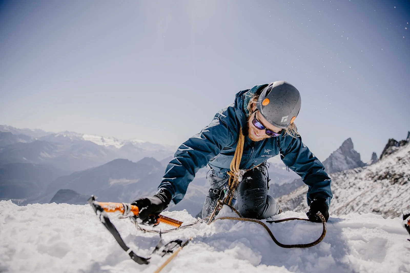 A man in winter gear and a helmet climbing snowy mountain terrain with a rope, against a backdrop of snow-covered mountains under a clear blue sky.