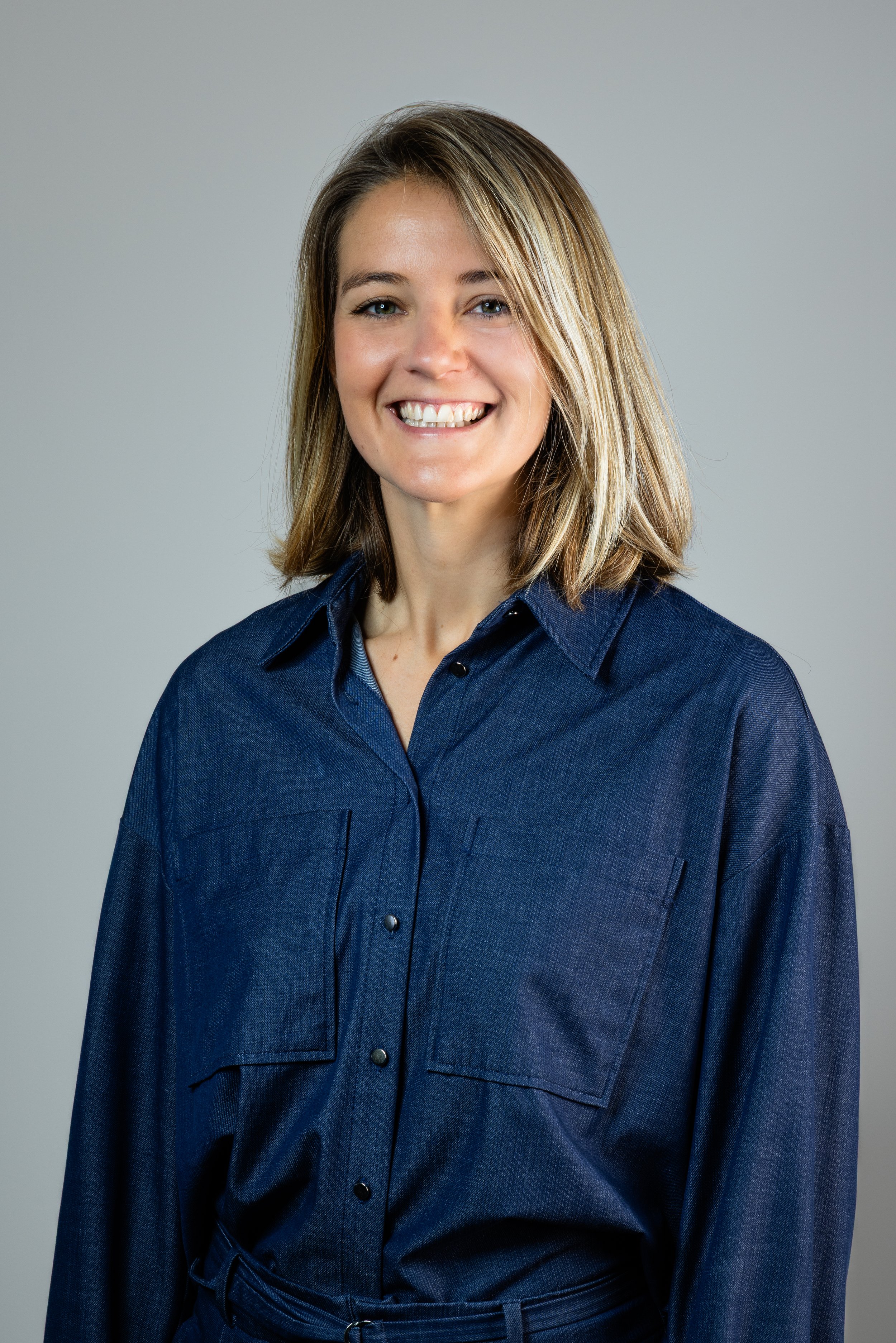 A smiling woman with shoulder-length blonde hair wearing a dark blue button-up shirt against a light gray background.