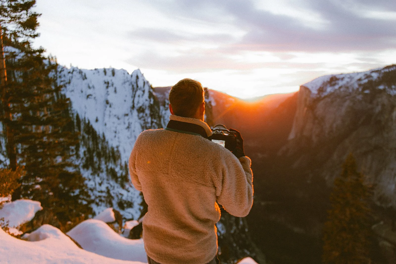 A person with a fleece jacket taking a photo with a camera during sunset in a snowy mountain landscape.