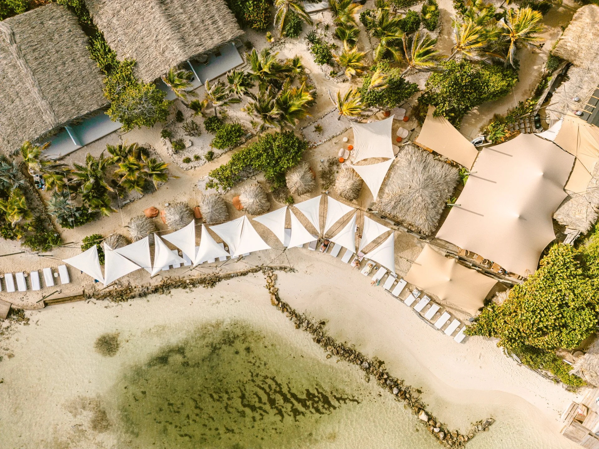 An aerial view of a tropical beach resort with sheared thatched roof buildings, white tents, lounge chairs, palm trees, green shrubbery, and a sandy shoreline with clear water.